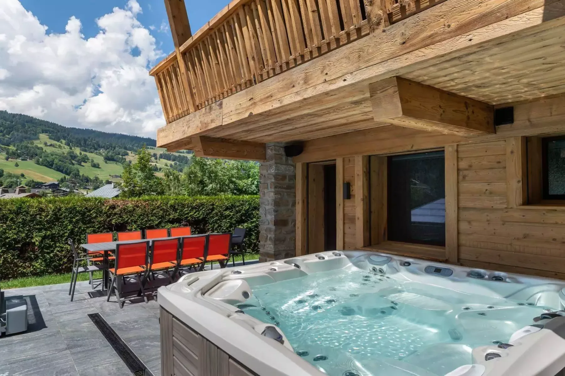 An outdoor hot tub beside a wooden chalet, with mountain scenery in the background.