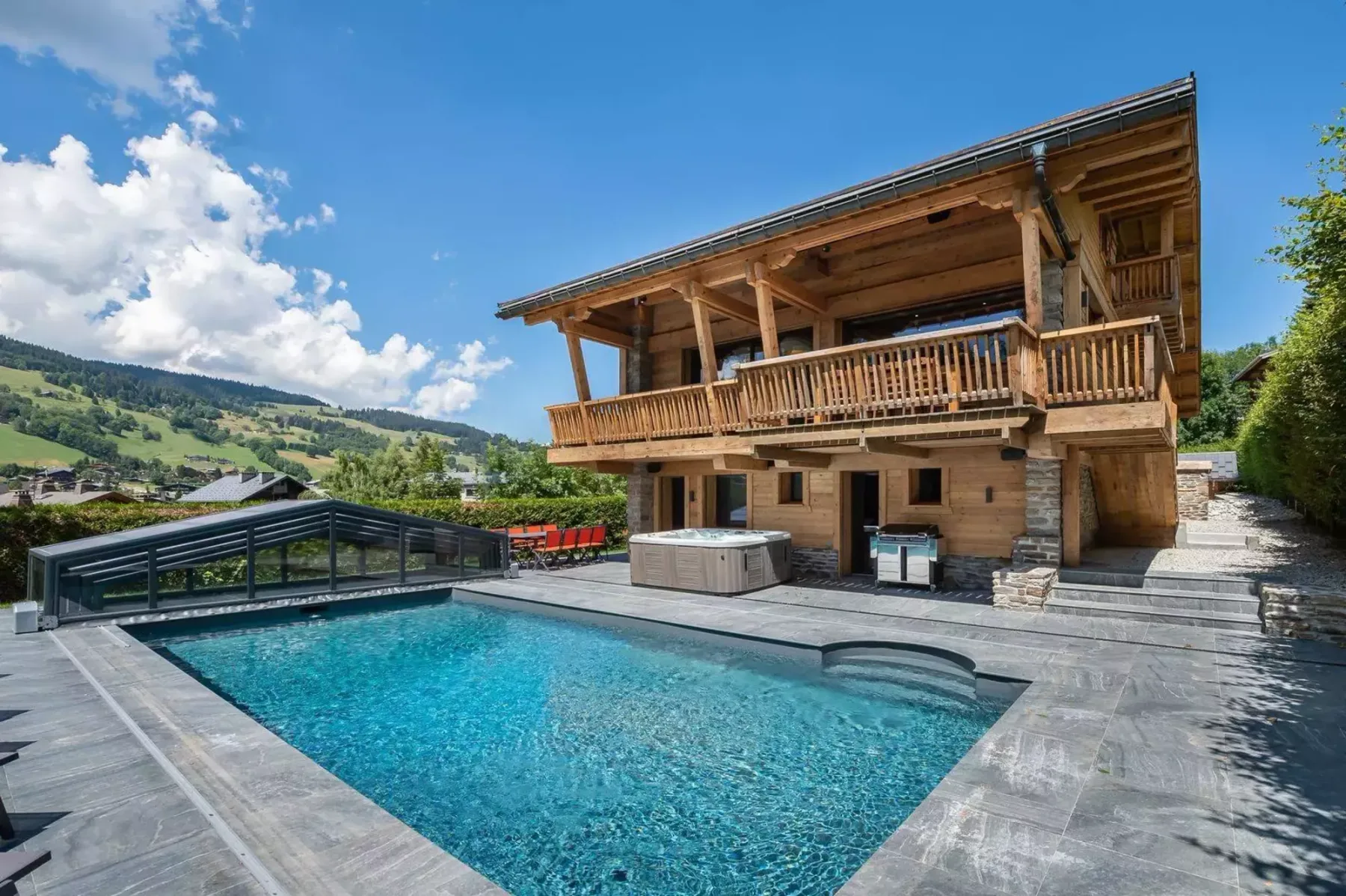 Un chalet con piscina y un amplio balcón de madera da a un paisaje montañoso verde.