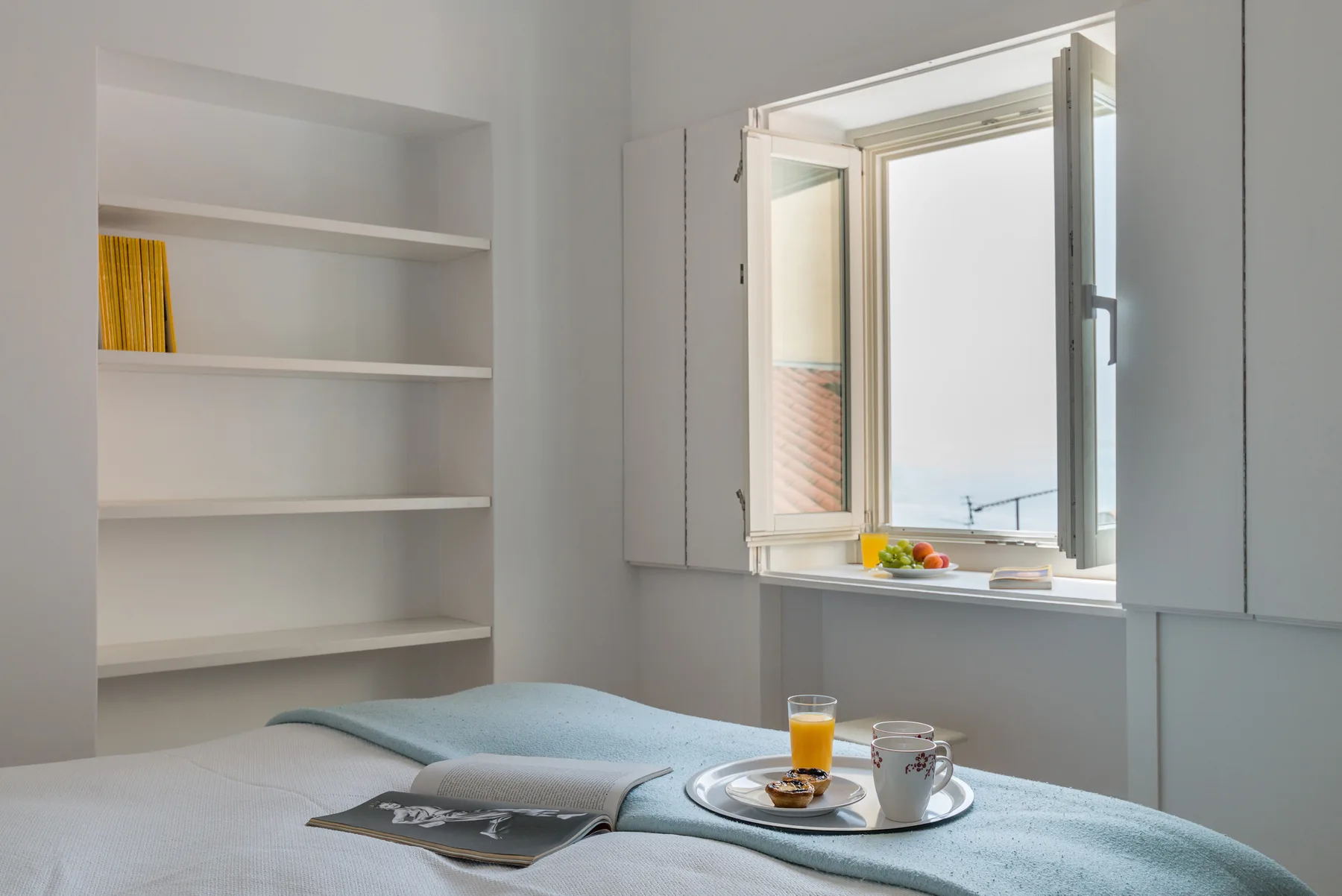 Un dormitorio con una cama junto a una ventana abierta y estantes empotrados.