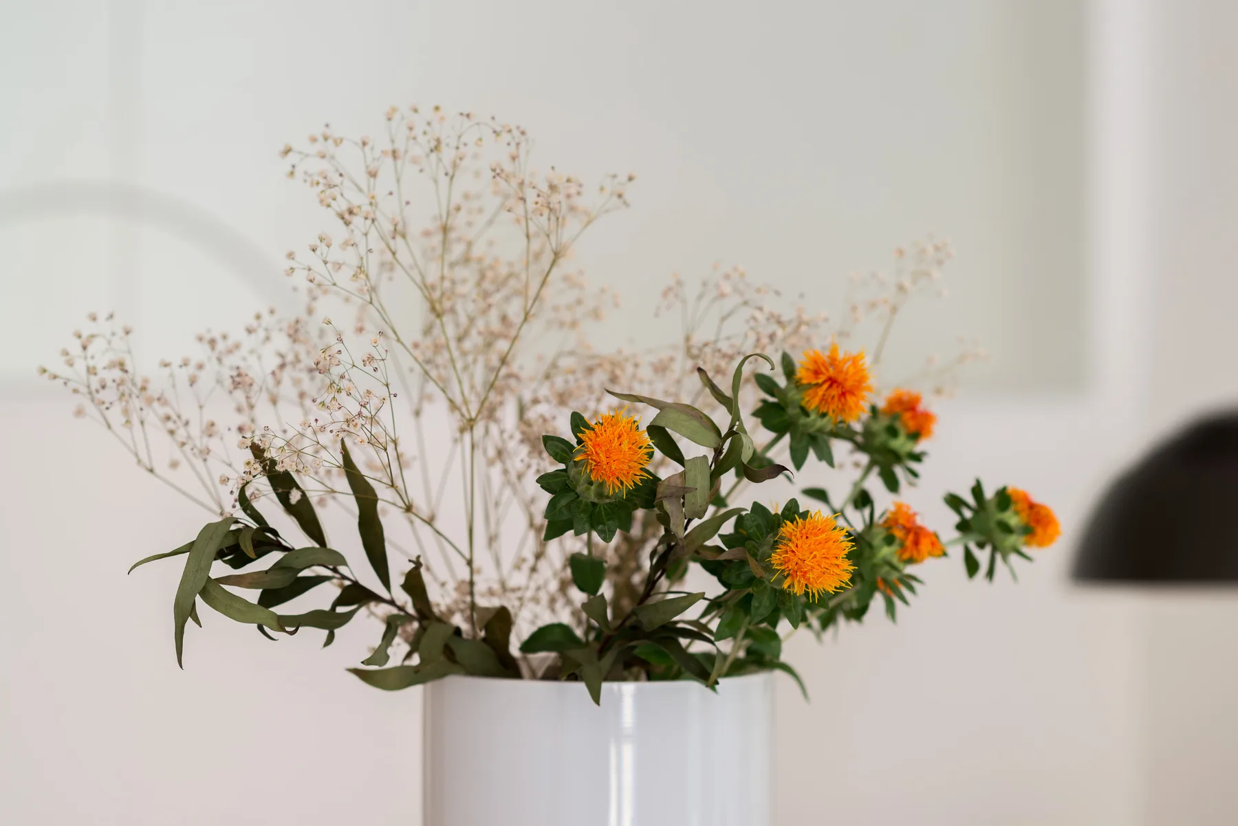 Un jarrón con flores naranjas y follaje está frente a una pared clara.