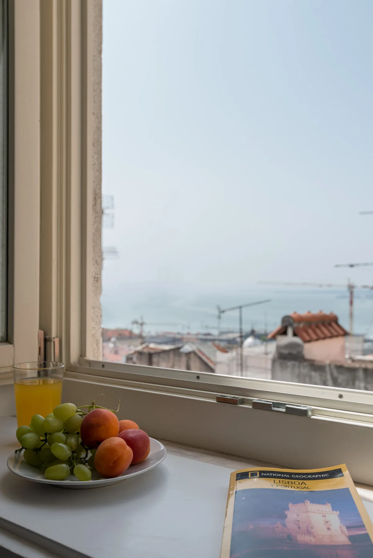Una ventana da a tejados y al agua, con fruta y un folleto en el alféizar.