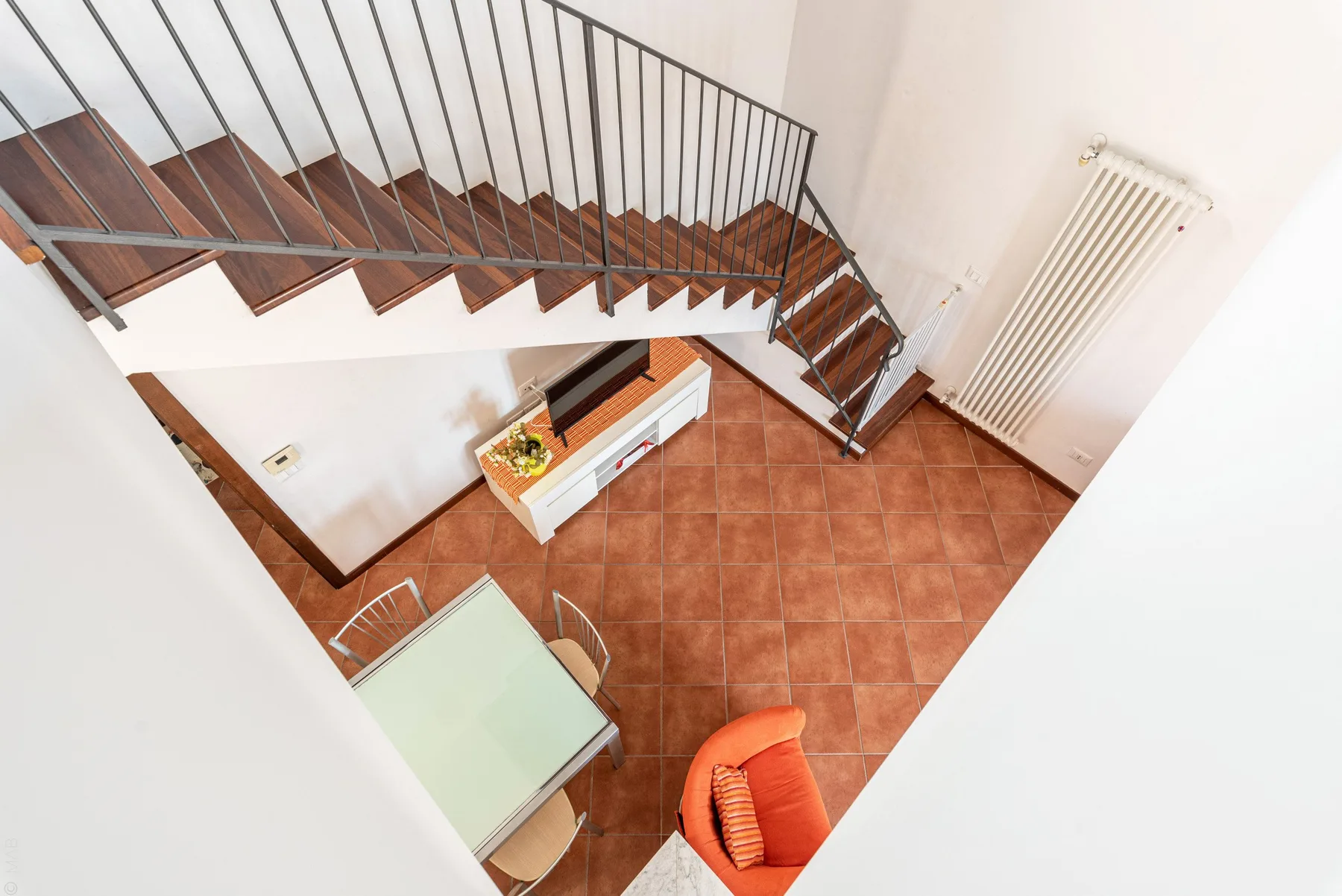 Un rellano interior con suelo de baldosas queda bajo una escalera, con una mesa y un radiador cerca.