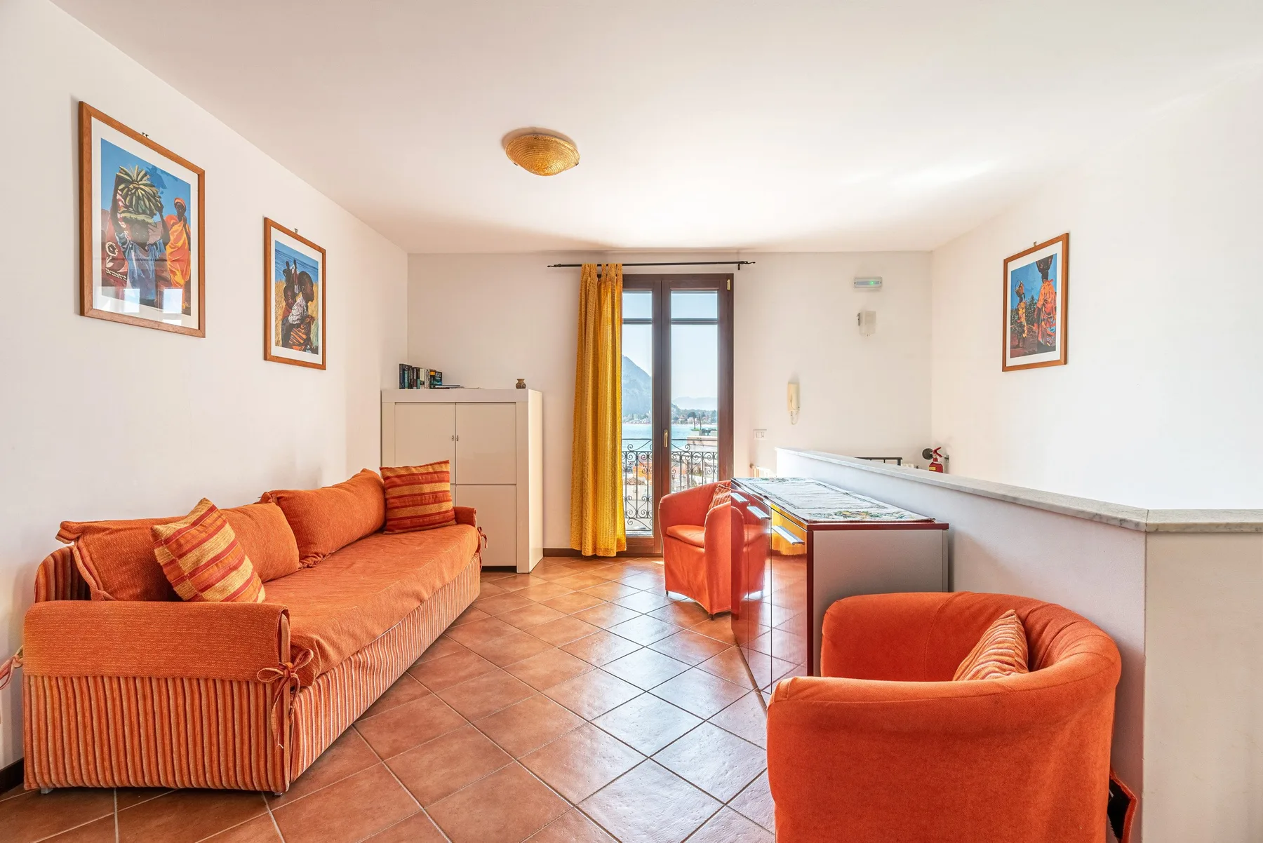 Una sala de estar luminosa con asientos naranjas, cuadros y una puerta de vidrio al balcón.