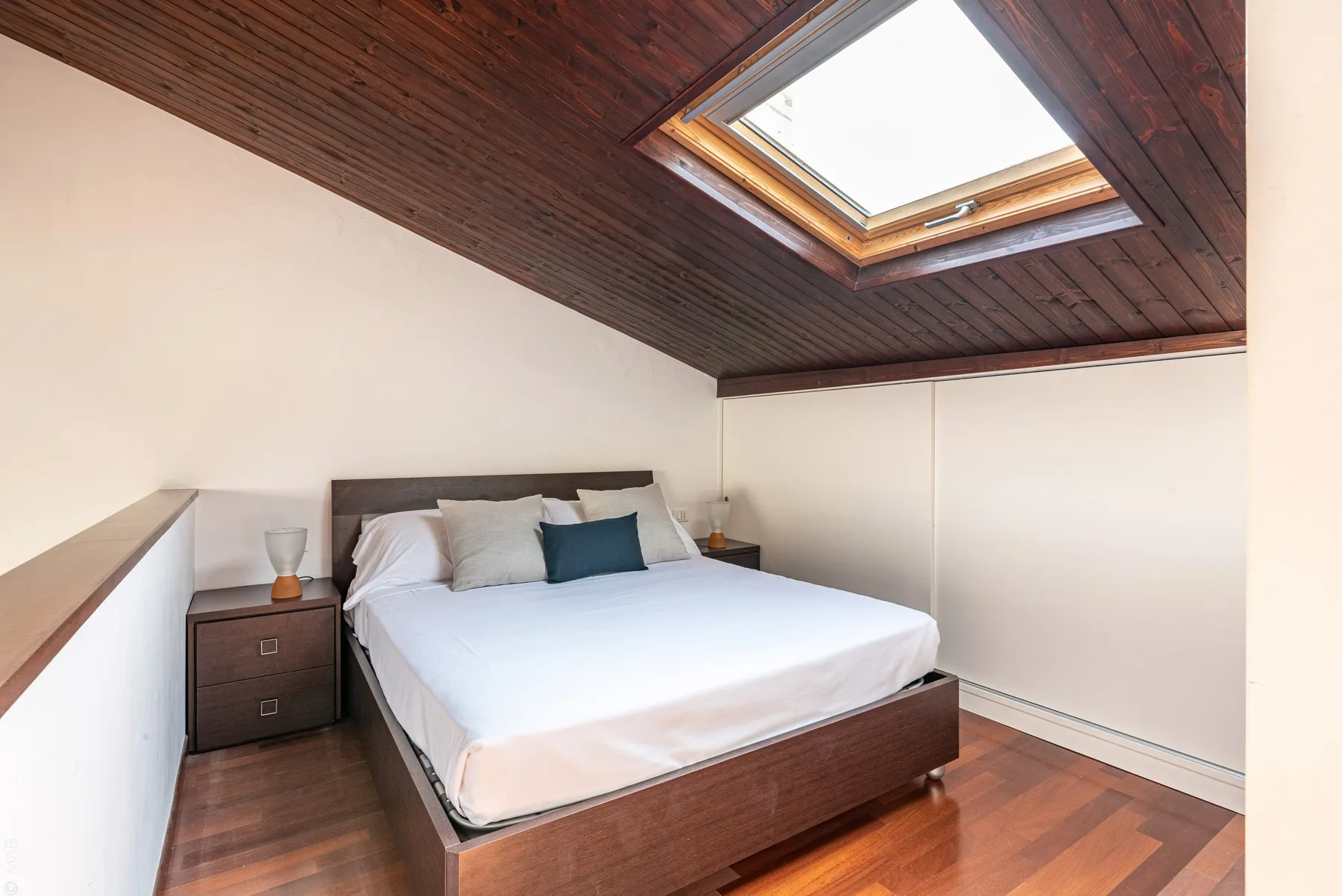 Un dormitorio con una cama bajo un techo inclinado de madera y una claraboya.