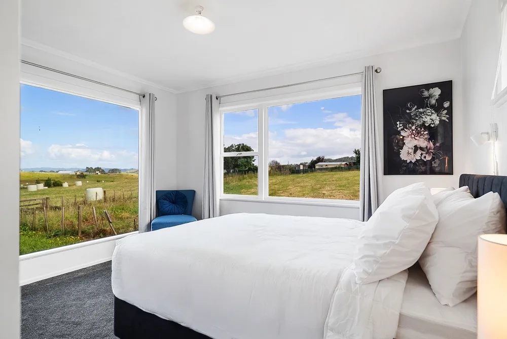 Dormitorio con cama blanca y grandes ventanas con vistas al campo.