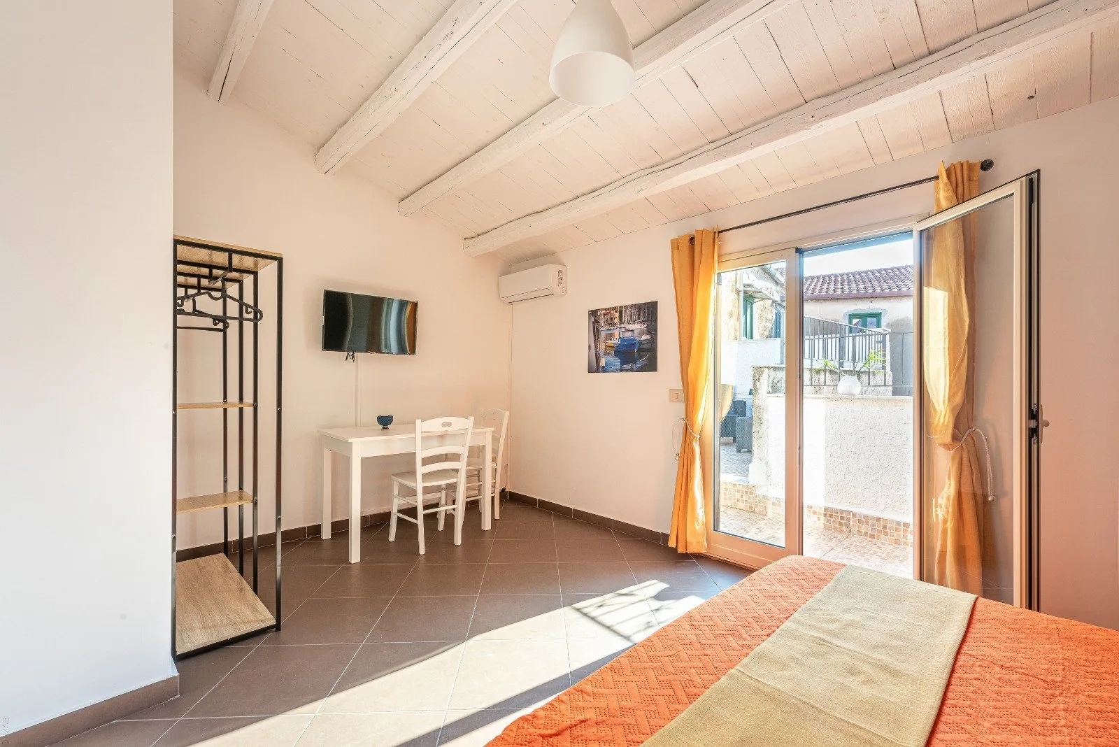 A bright bedroom with a bed, a small desk, and an open glass door to a terrace.
