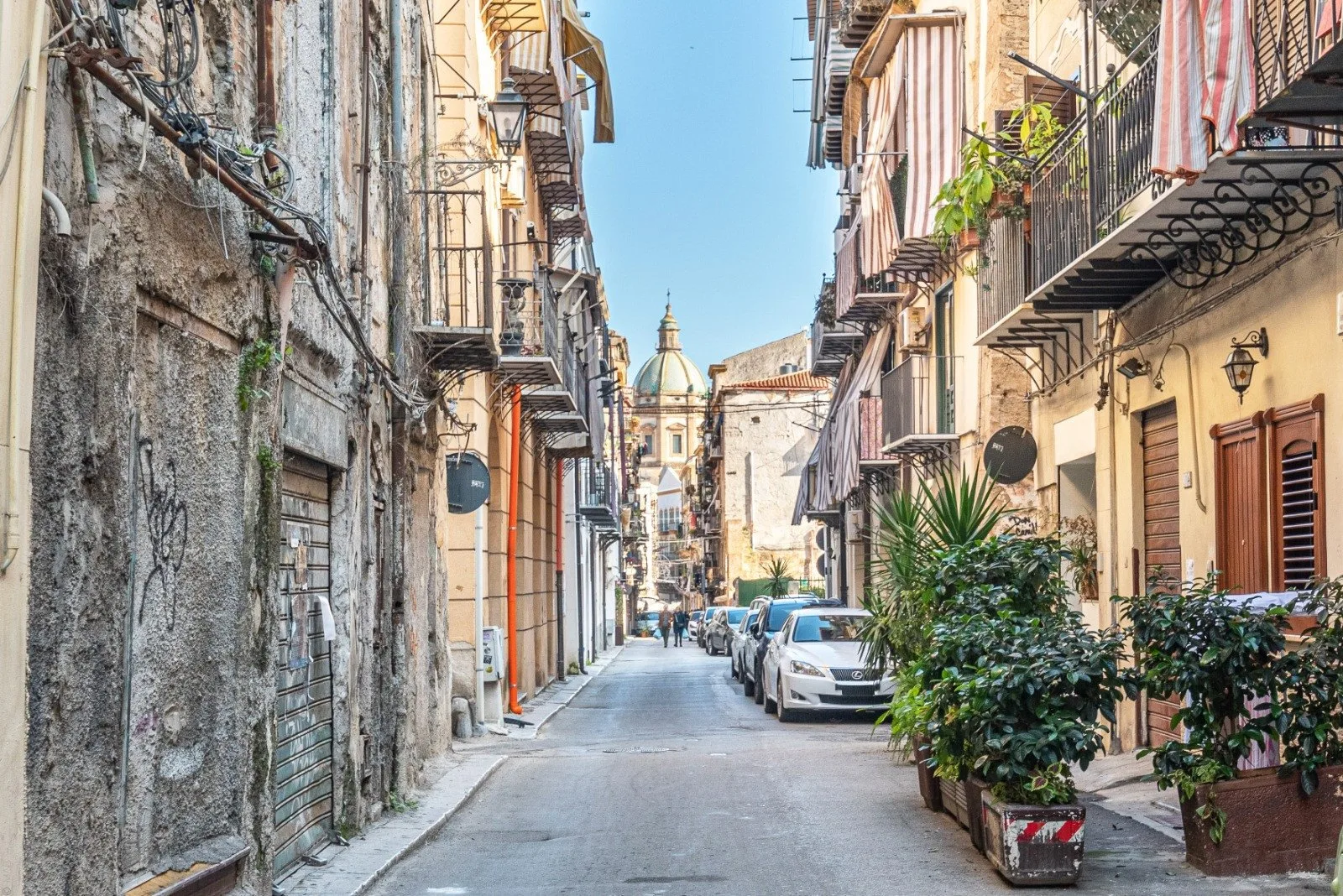 A narrow city street lined with buildings, parked cars, and potted plants.