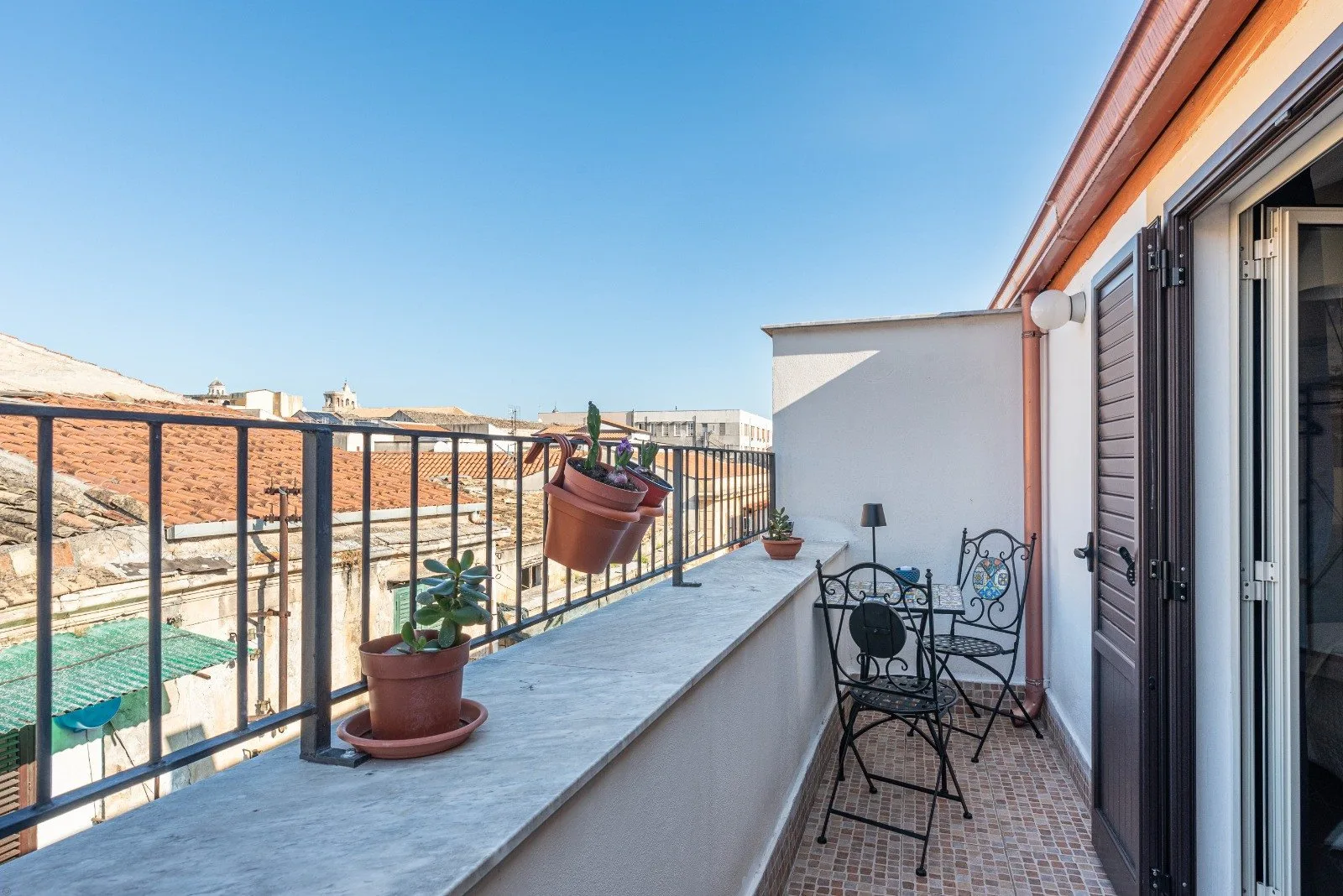 A narrow balcony with outdoor seating and potted plants overlooks nearby rooftops.