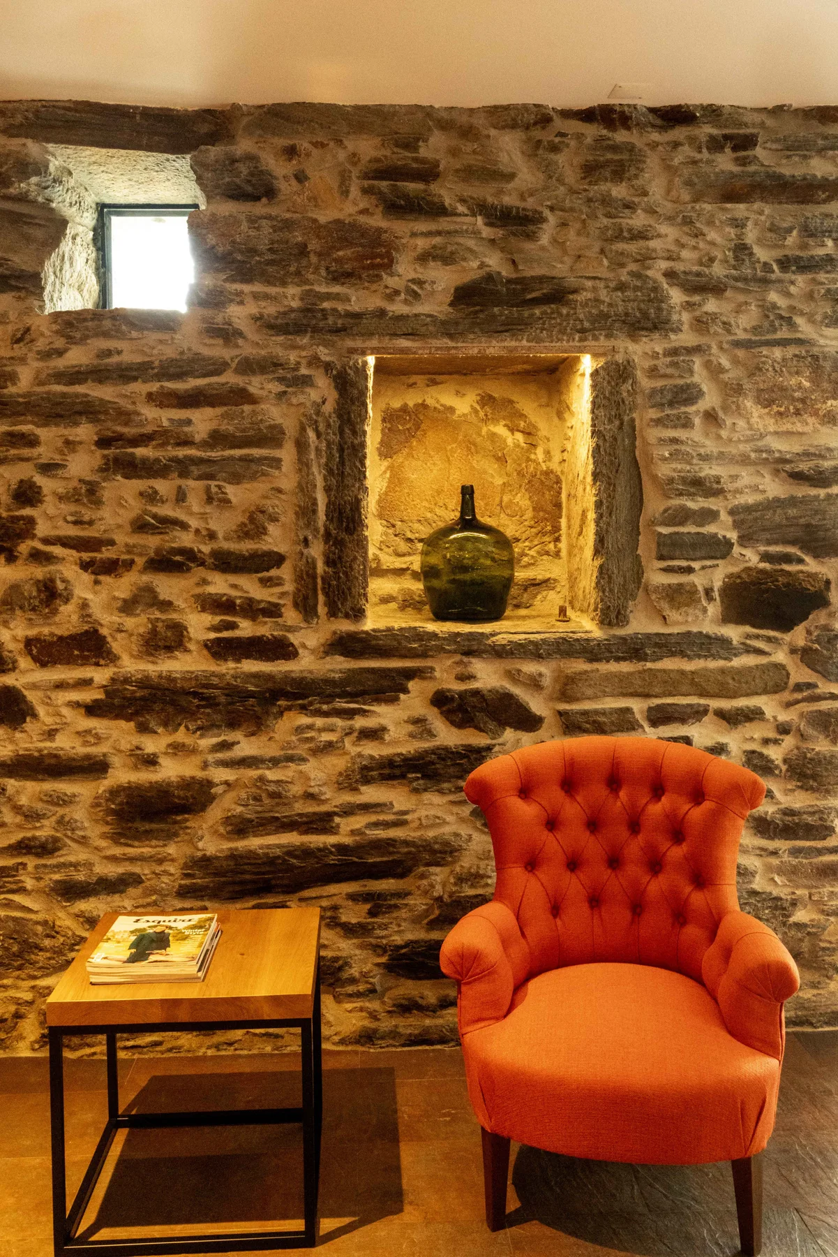 Un sillón naranja está junto a una mesa pequeña frente a una pared de piedra.