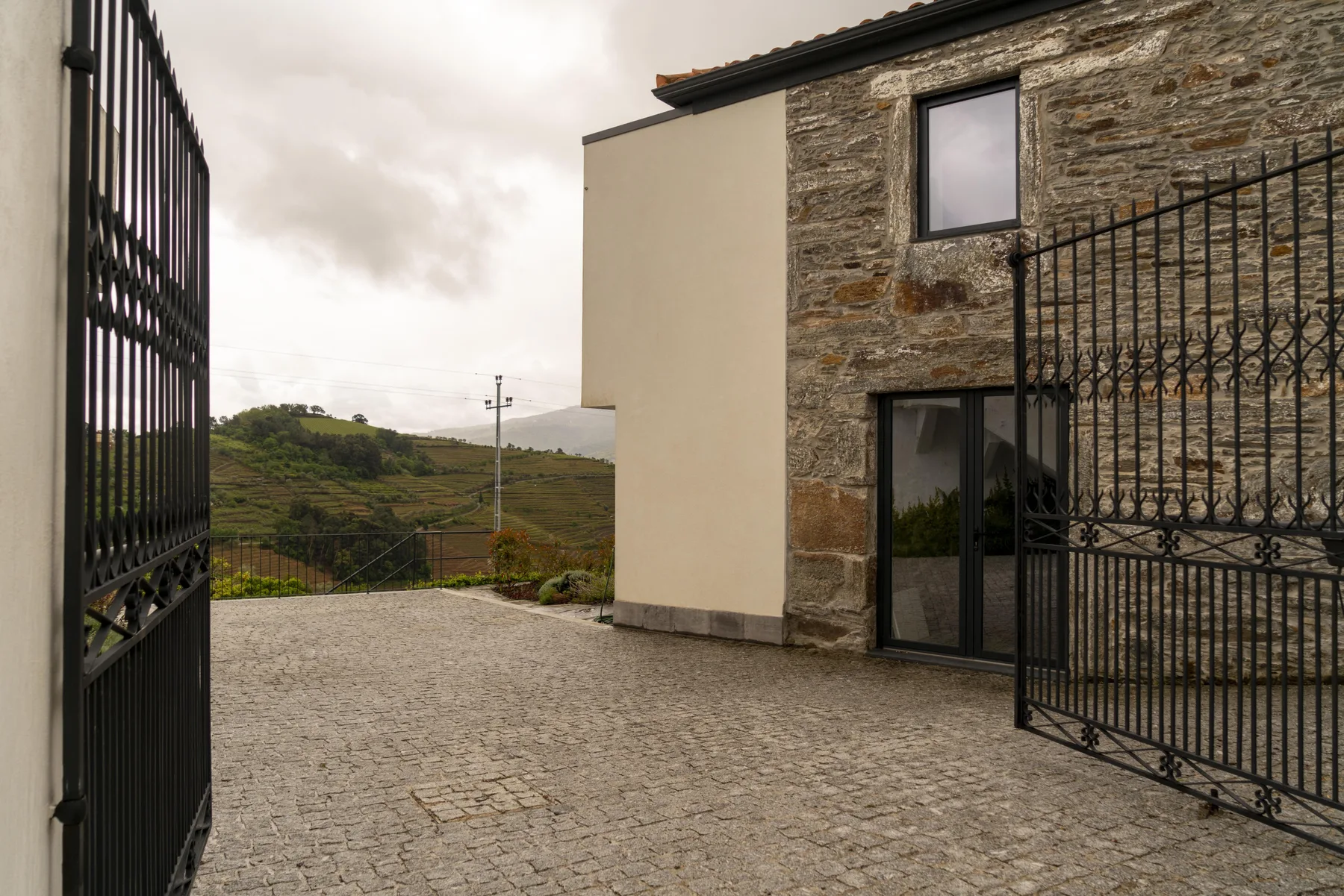 Un patio empedrado junto a un edificio de piedra y yeso, con las puertas abiertas y colinas al fondo.