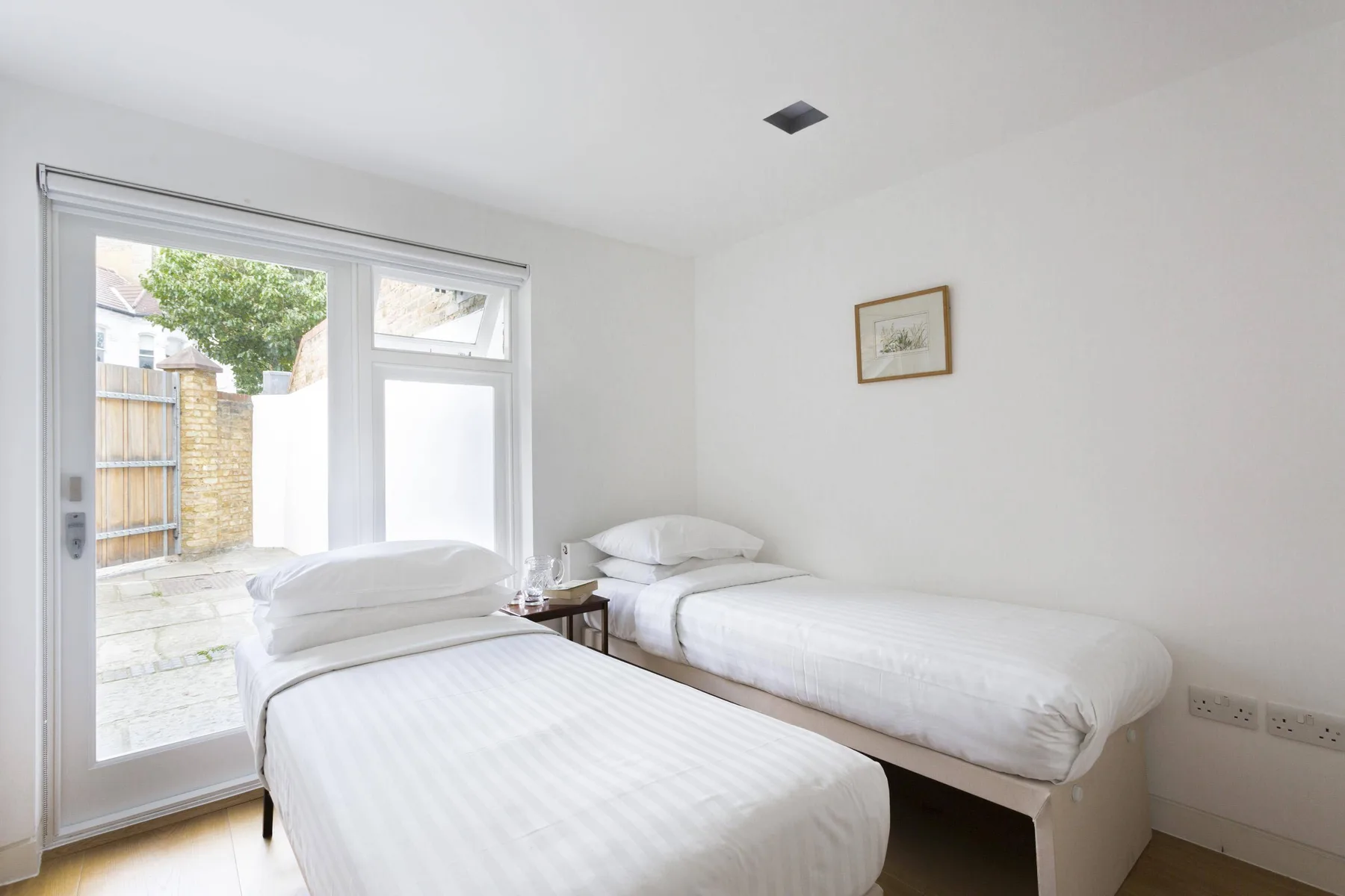 A bright bedroom with two beds beside a large window opening onto a patio.