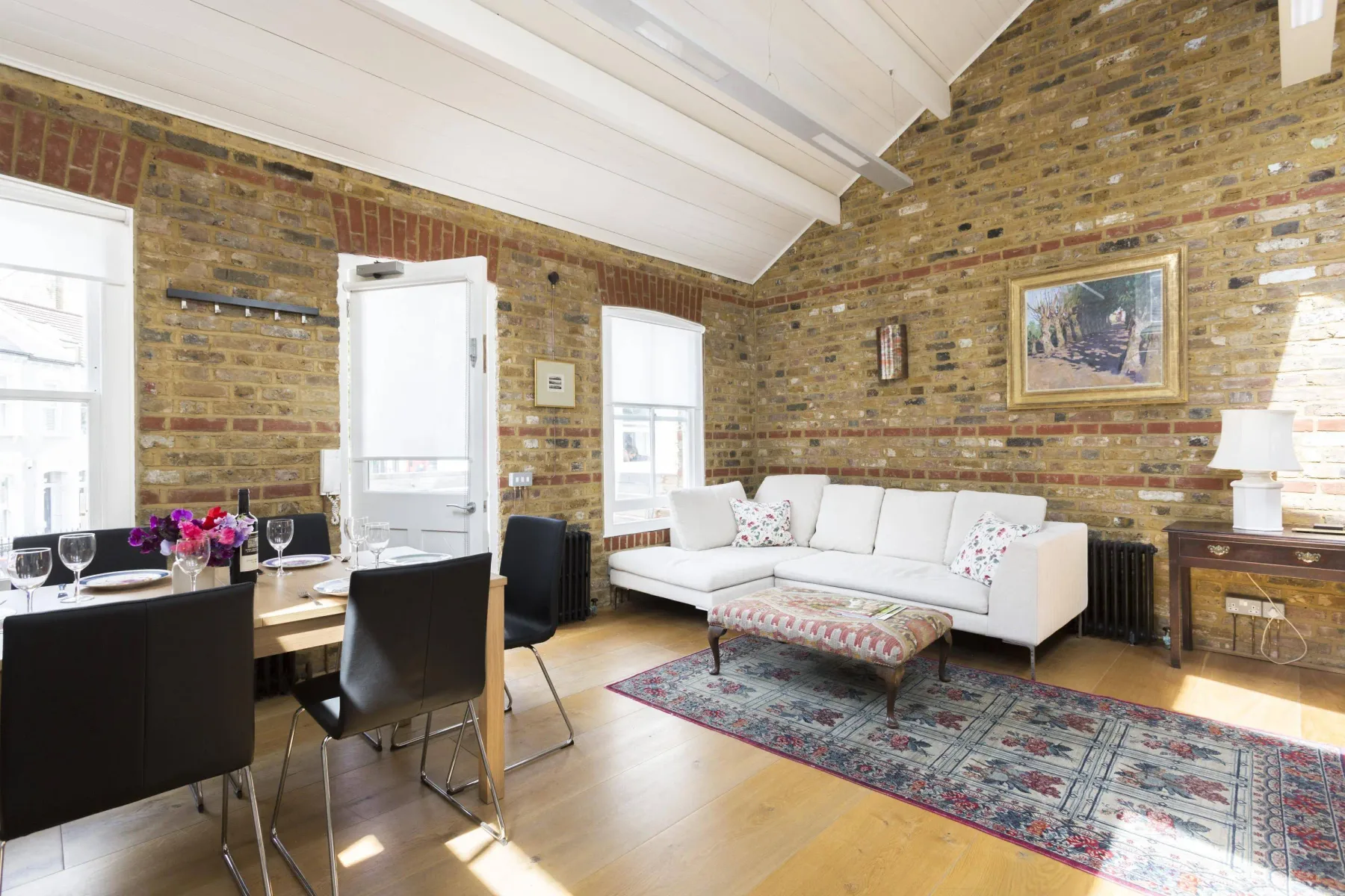 A living room with white seating and a dining table beside exposed brick walls.