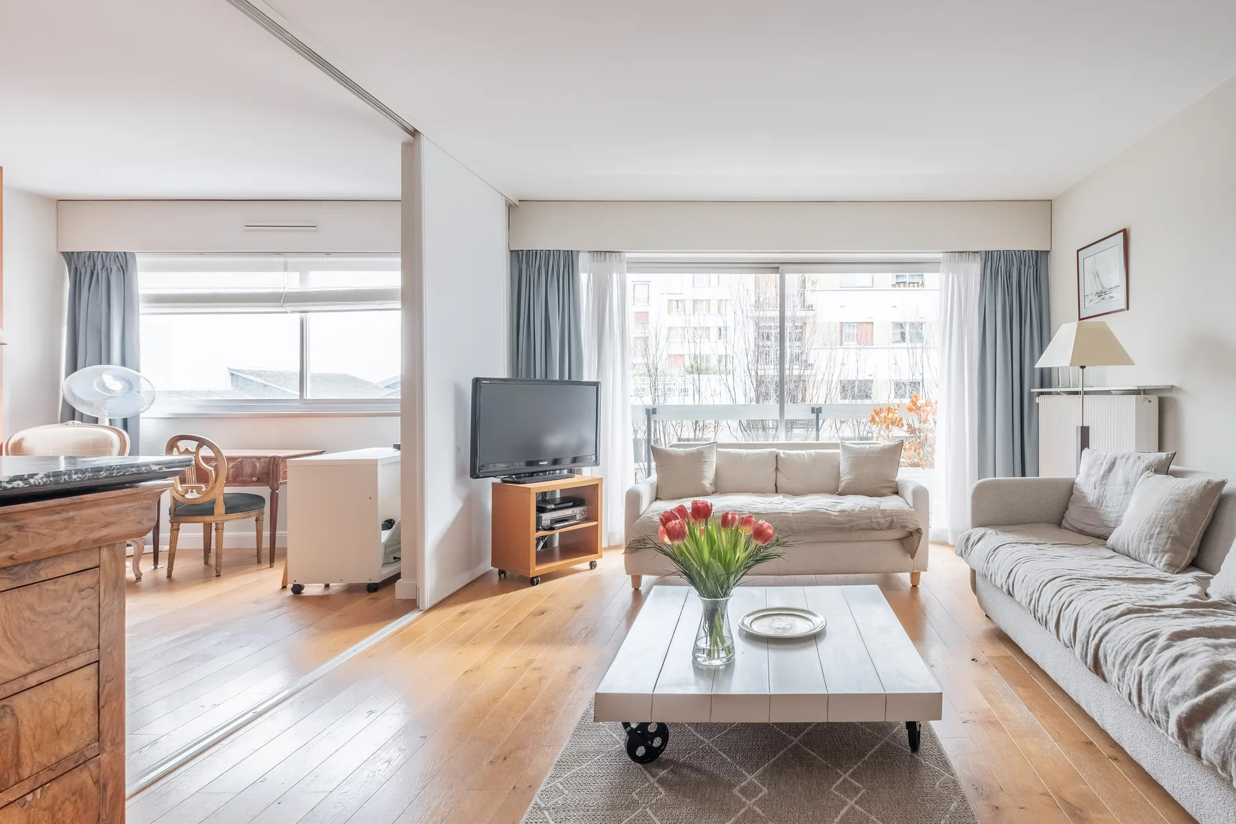 A bright living room with seating, a television, and a dining area by the windows.