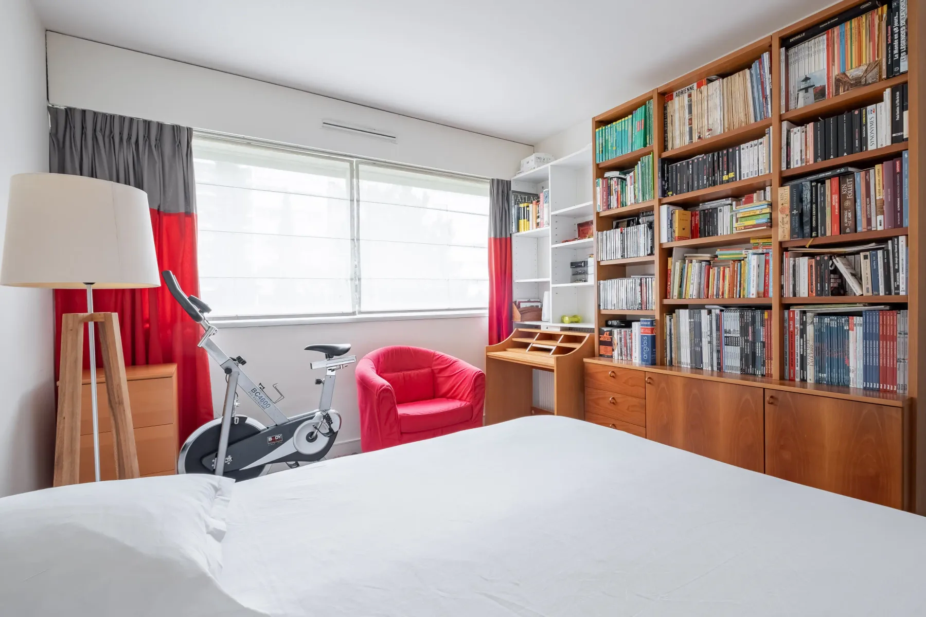 A bedroom with a large bookcase, exercise bike, and red armchair.