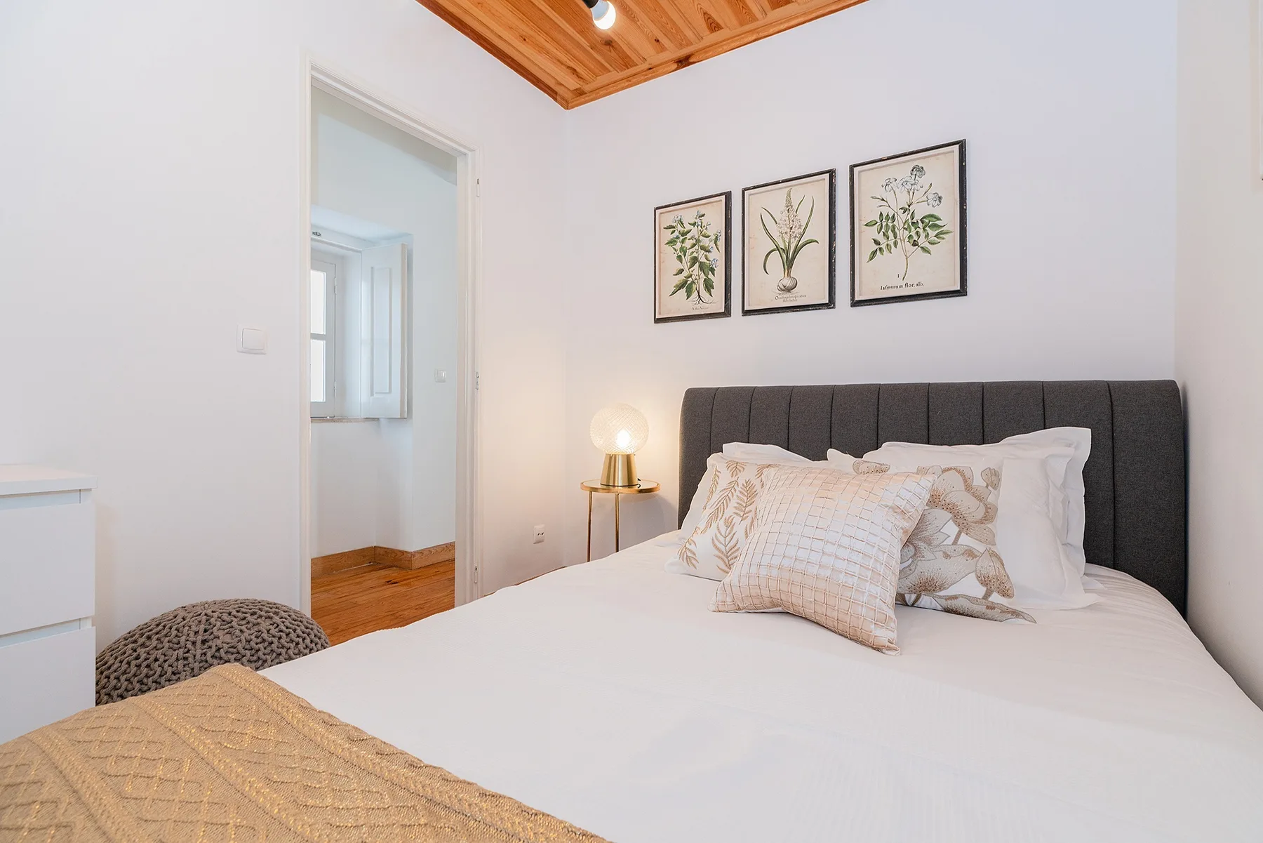 A bedroom with a bed, framed botanical prints, and a wood-paneled ceiling.