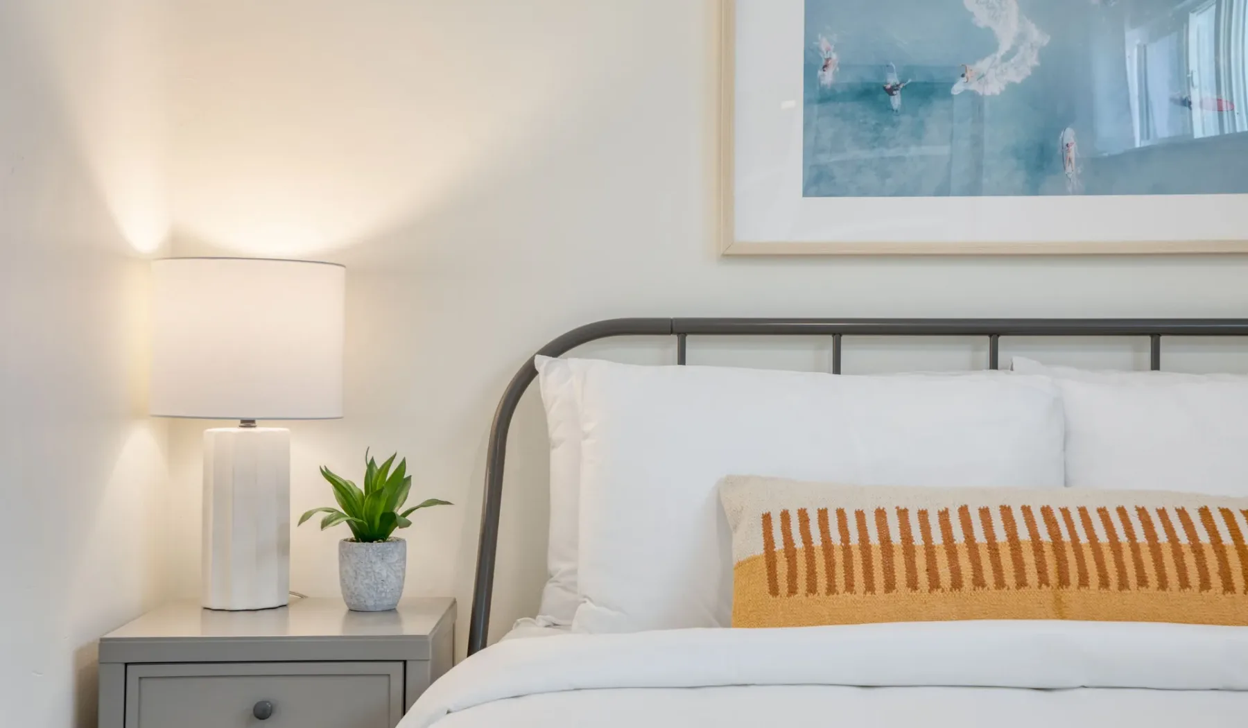 Un dormitorio con una cama bien tendida, una lámpara de mesita y una planta pequeña.