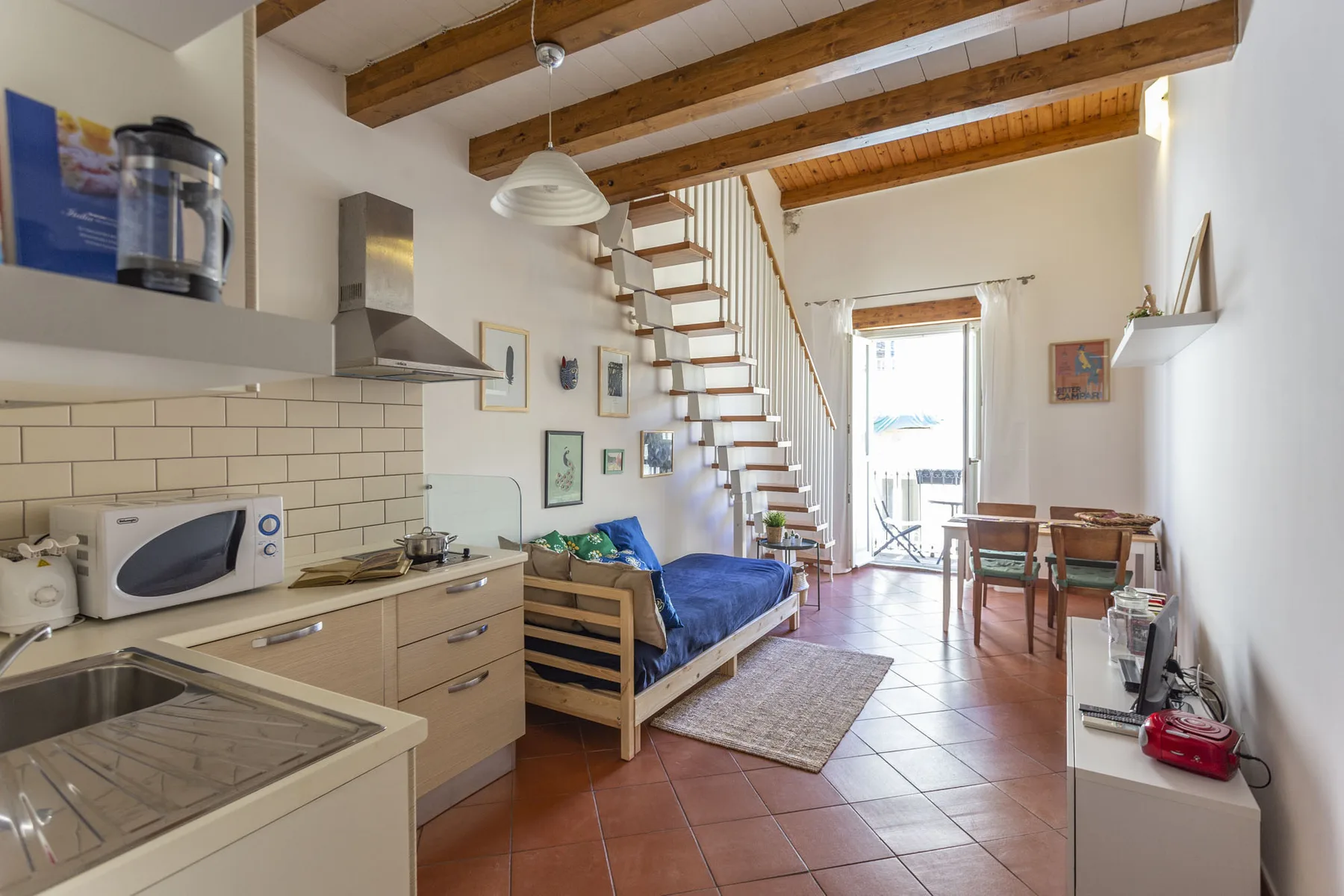 A compact kitchen and dining area share an open room with a bed and staircase.