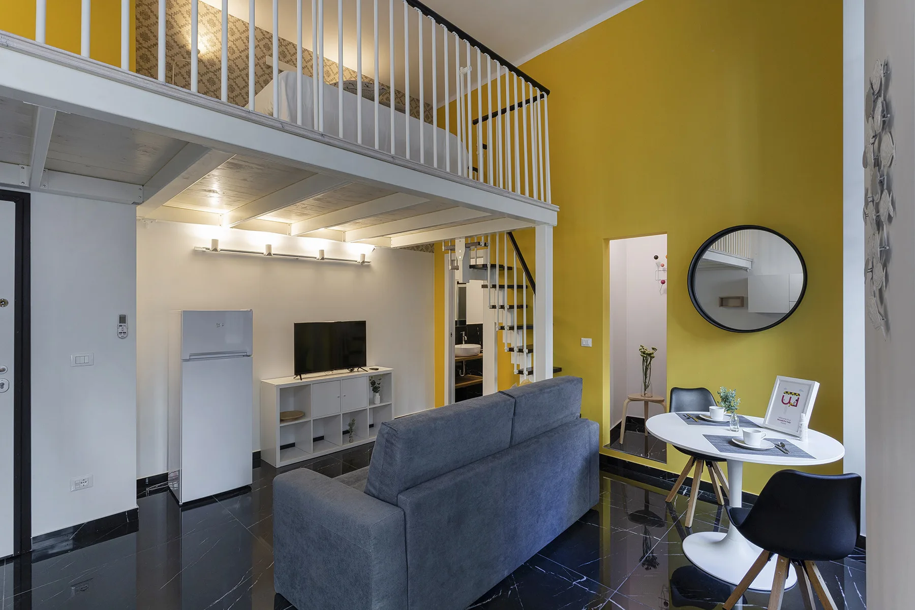 Una sala compacta con sofá gris, mesa de comedor y dormitorio elevado.