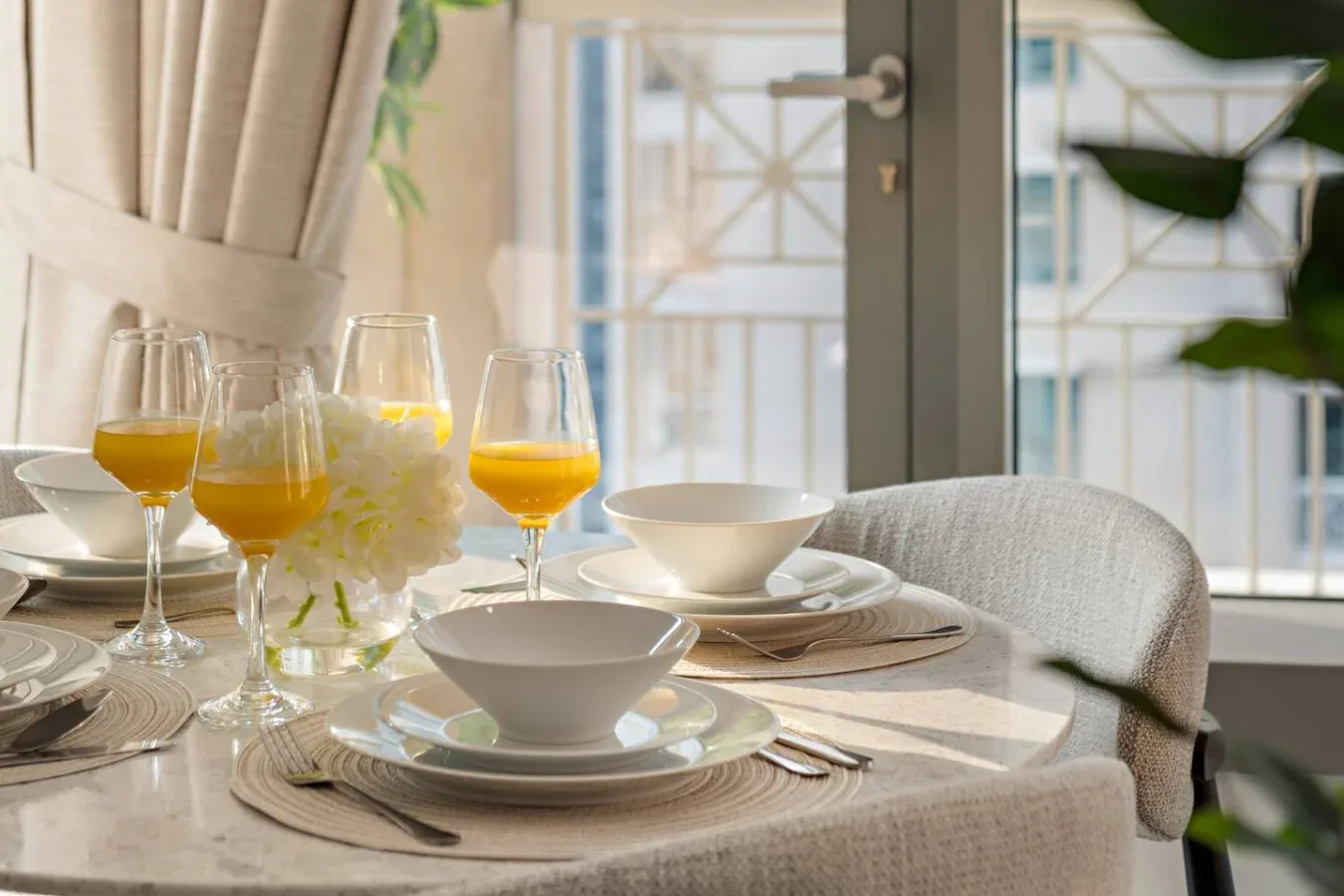 Una mesa de comedor está puesta con platos y copas junto a una ventana luminosa.