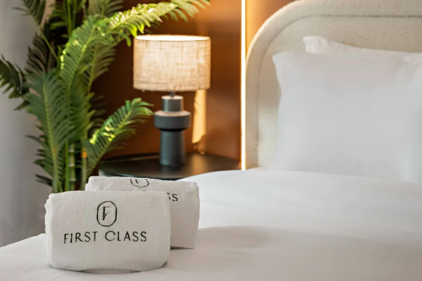 Un dormitorio con ropa de cama blanca, una lámpara de noche y plantas frondosas.