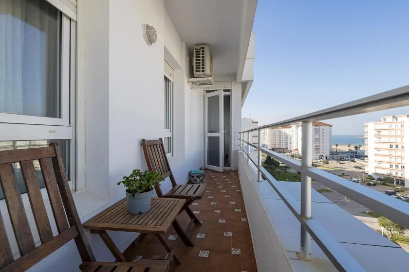 A narrow balcony with wooden seating overlooks nearby buildings and the sea.