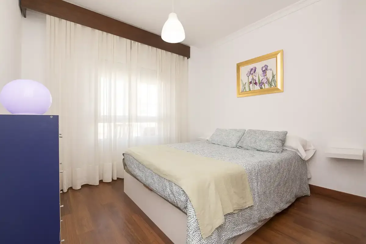 A bedroom with a bed, sheer curtains, and a framed floral picture.