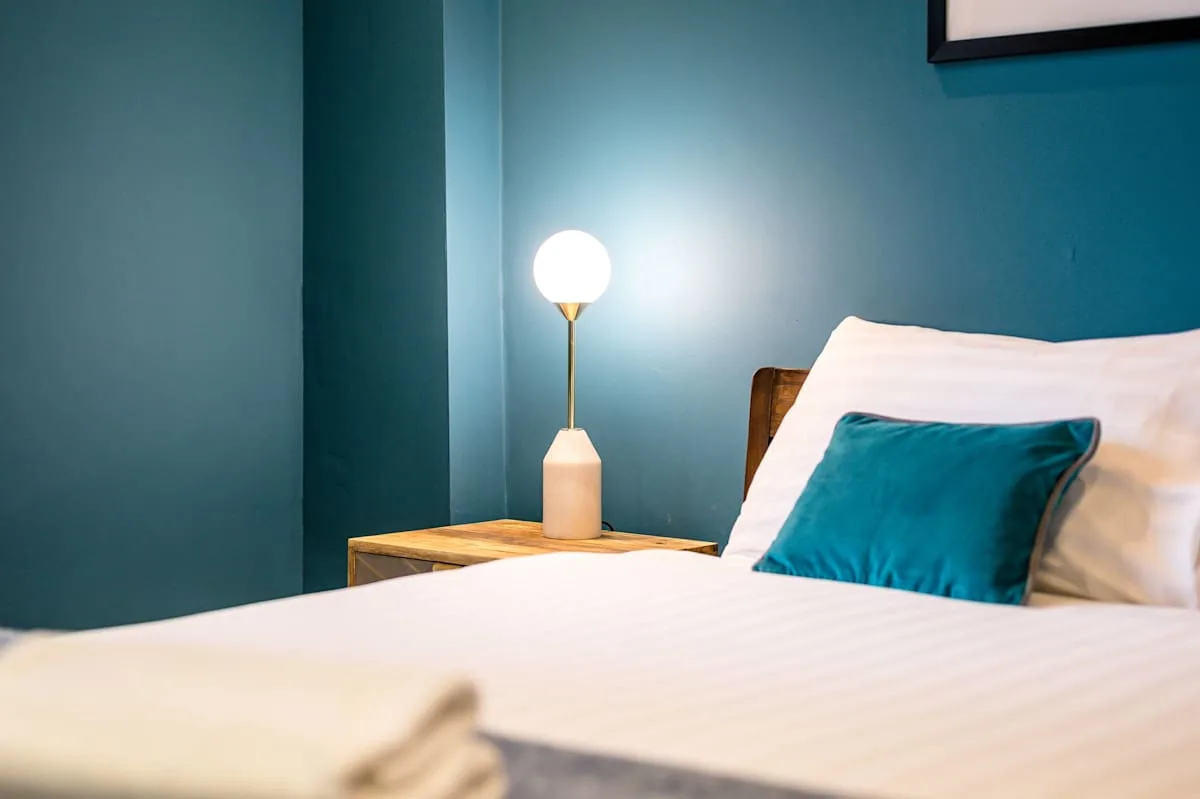 A bedroom with white bedding and a teal accent pillow beside a lit lamp.