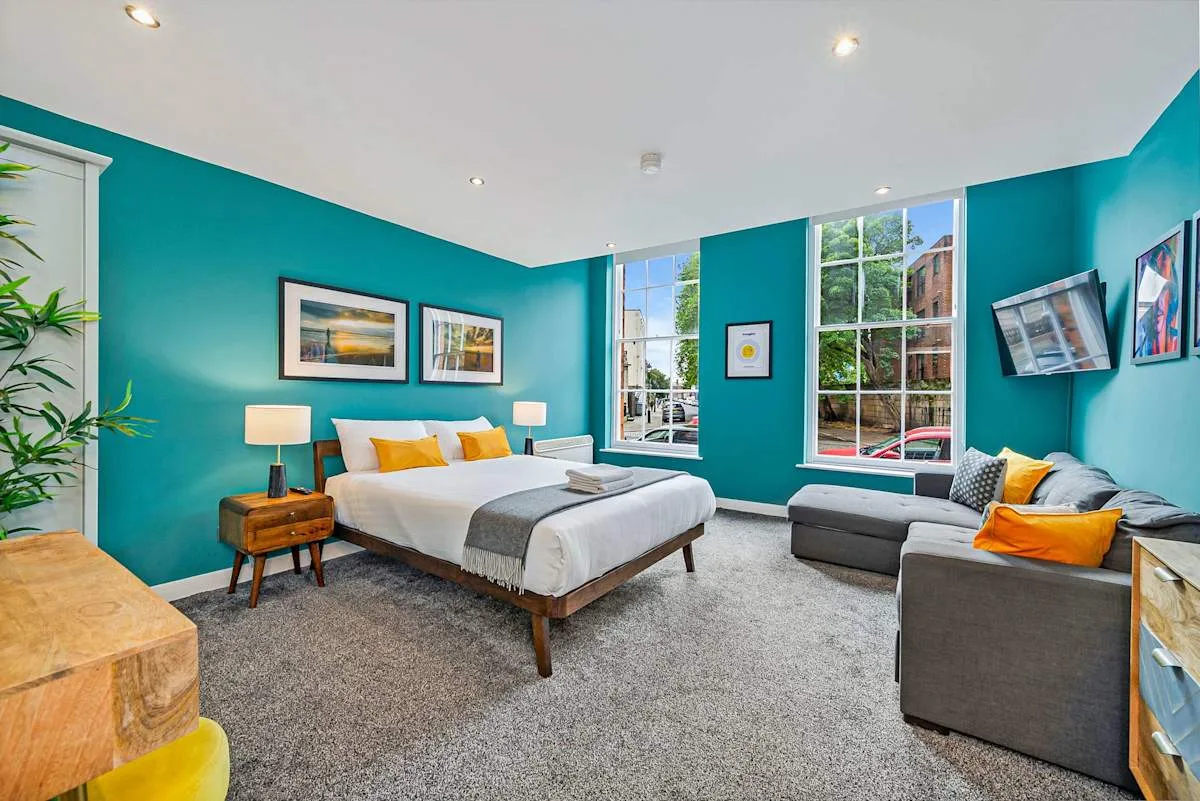 A bedroom with a bed and seating area, framed by teal walls and large windows.