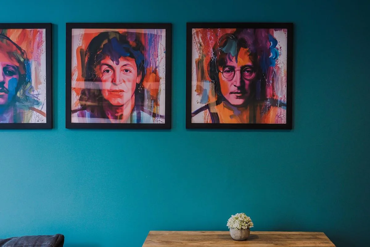 A teal-walled room displays colorful framed portraits above a wooden table.