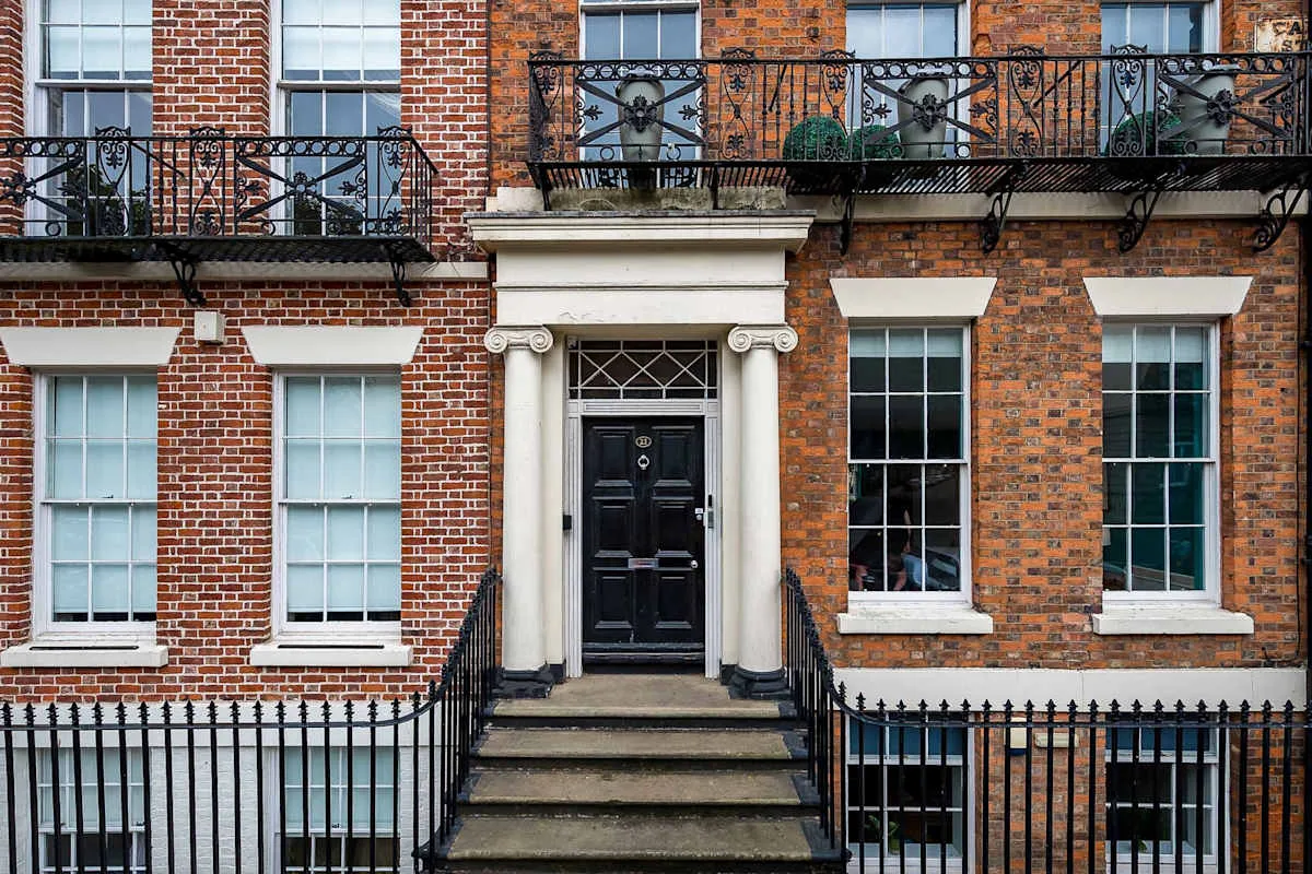 A brick townhouse facade has a black front door, stone steps, and wrought-iron railings.