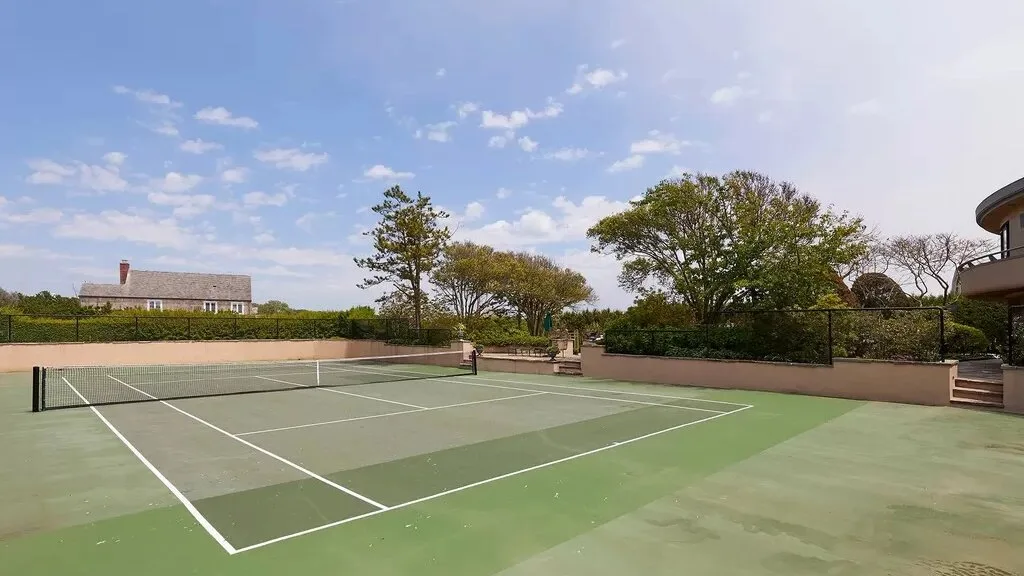 An outdoor tennis court with trees and a low wall beyond it.
