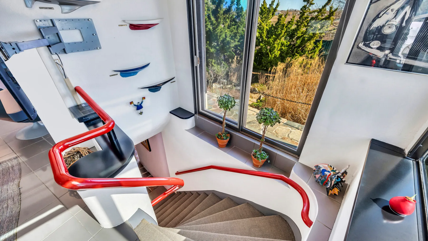 A curved staircase descends beside large windows overlooking trees and greenery.