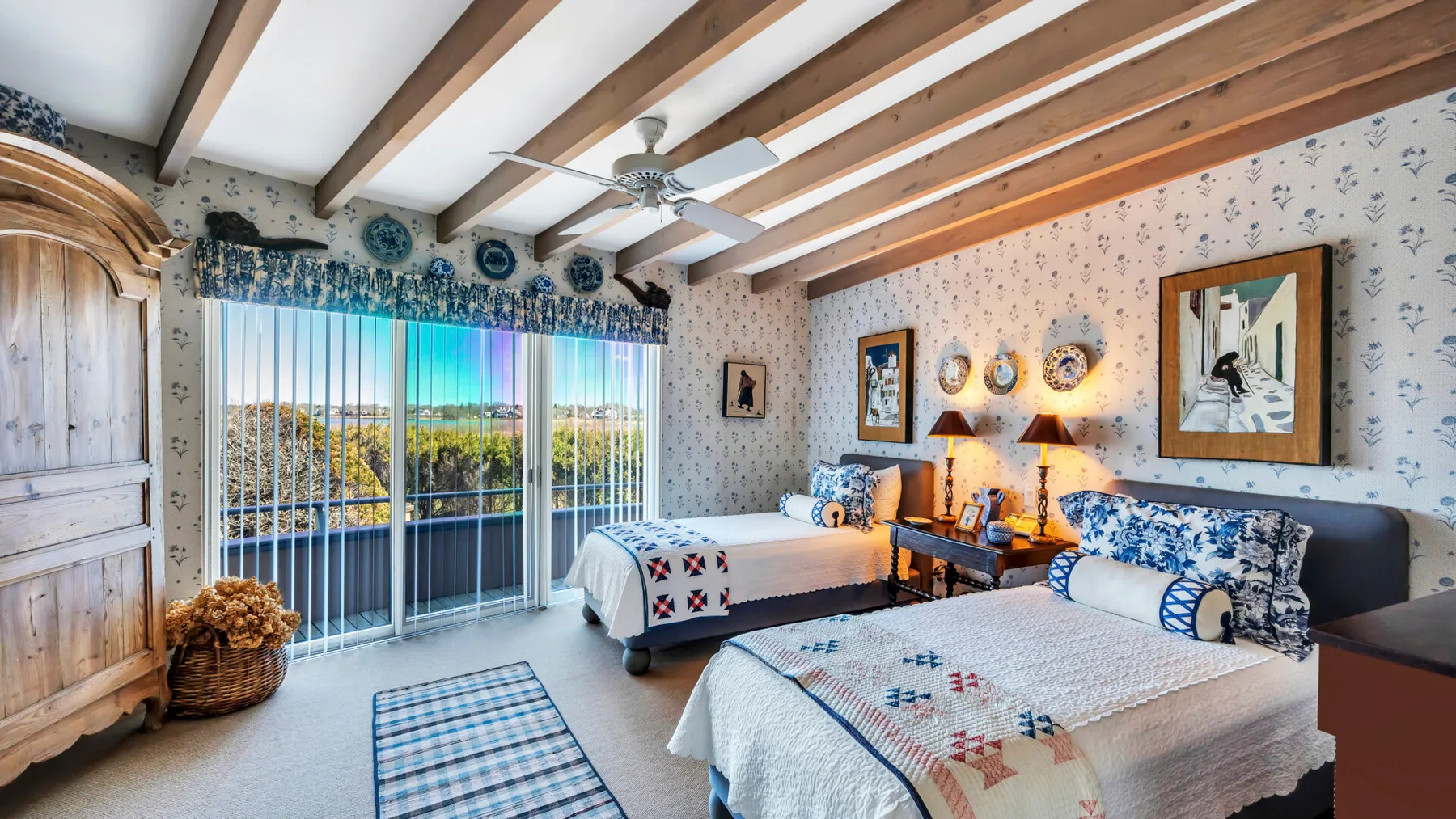 A bedroom with two beds, patterned bedding, and large windows beneath exposed ceiling beams.