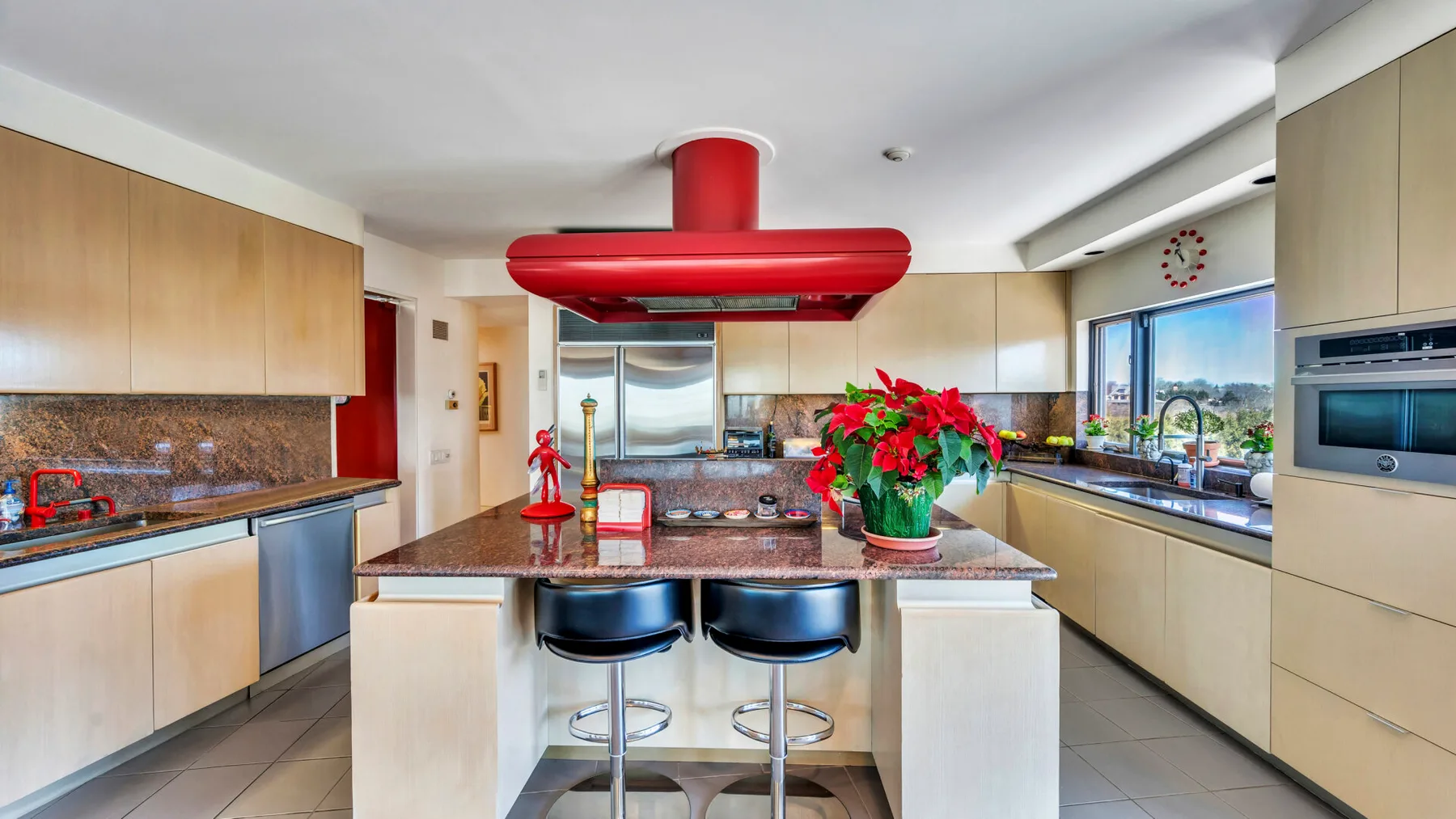 A modern kitchen with a central island, red range hood, and windows along the counter.
