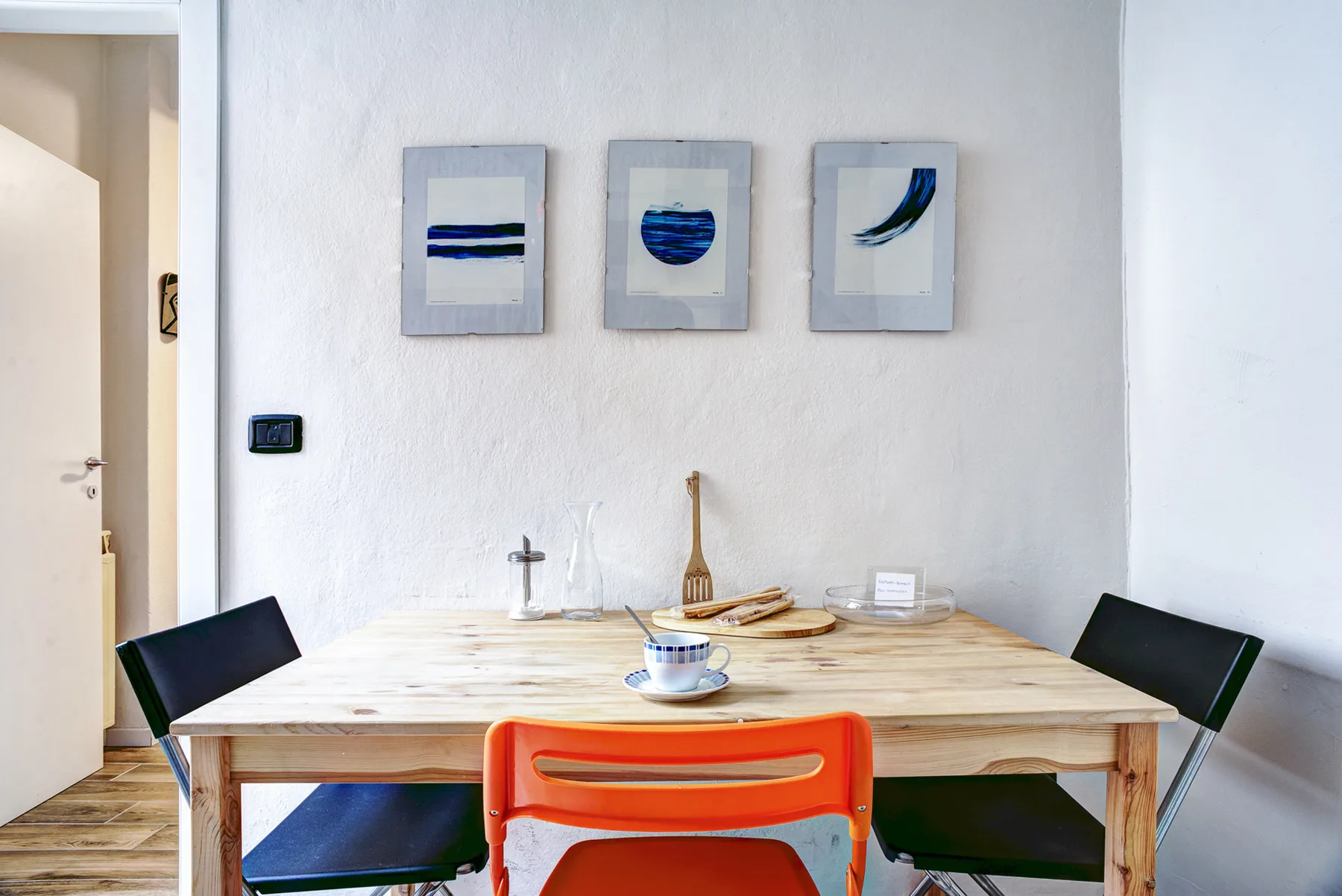 A dining table sits beneath three framed artworks, with colorful chairs around it.