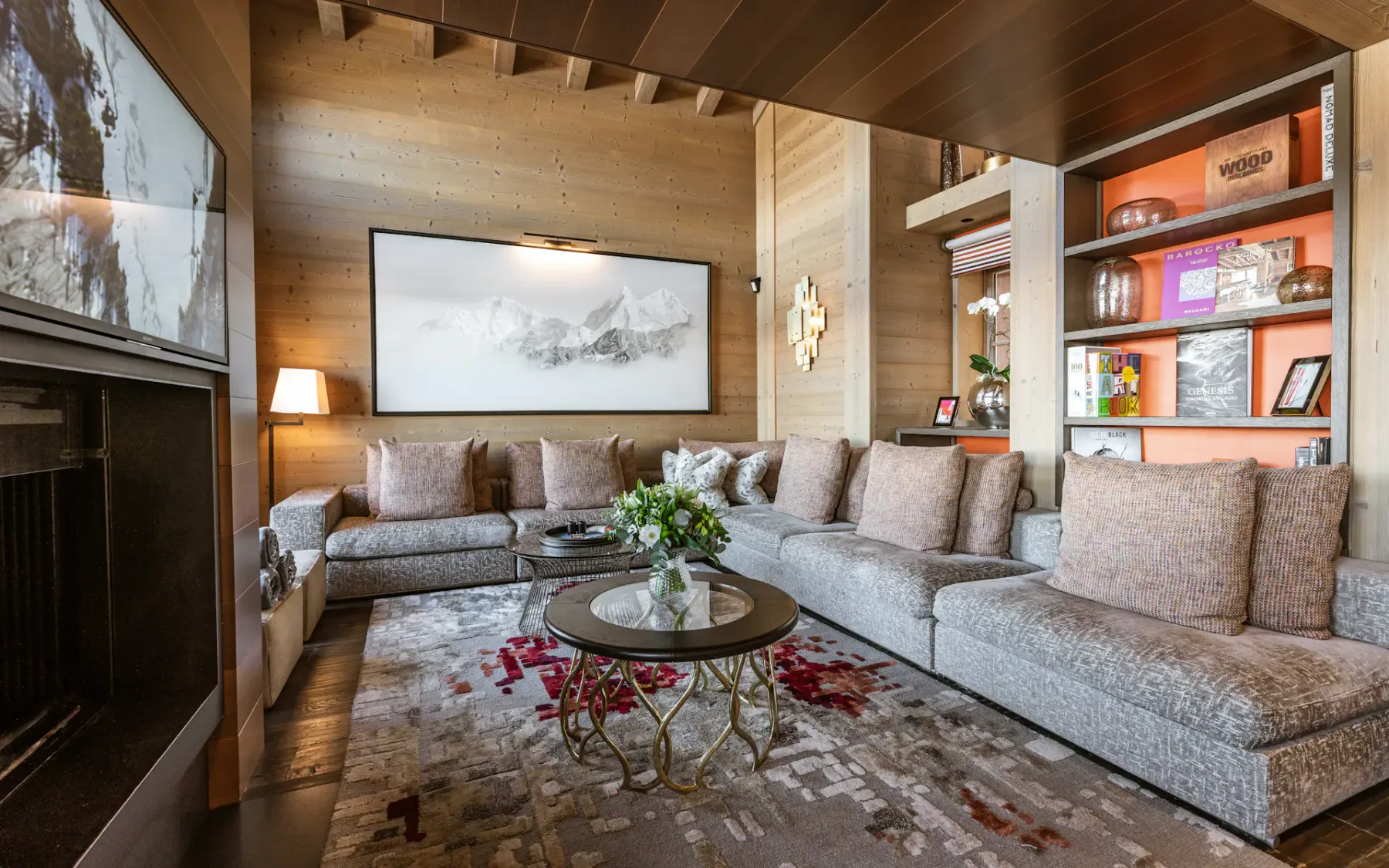 Cozy living room with a large sectional sofa, abstract art, a round coffee table, bookshelves, and a patterned rug. Warm lighting enhances the ambiance.