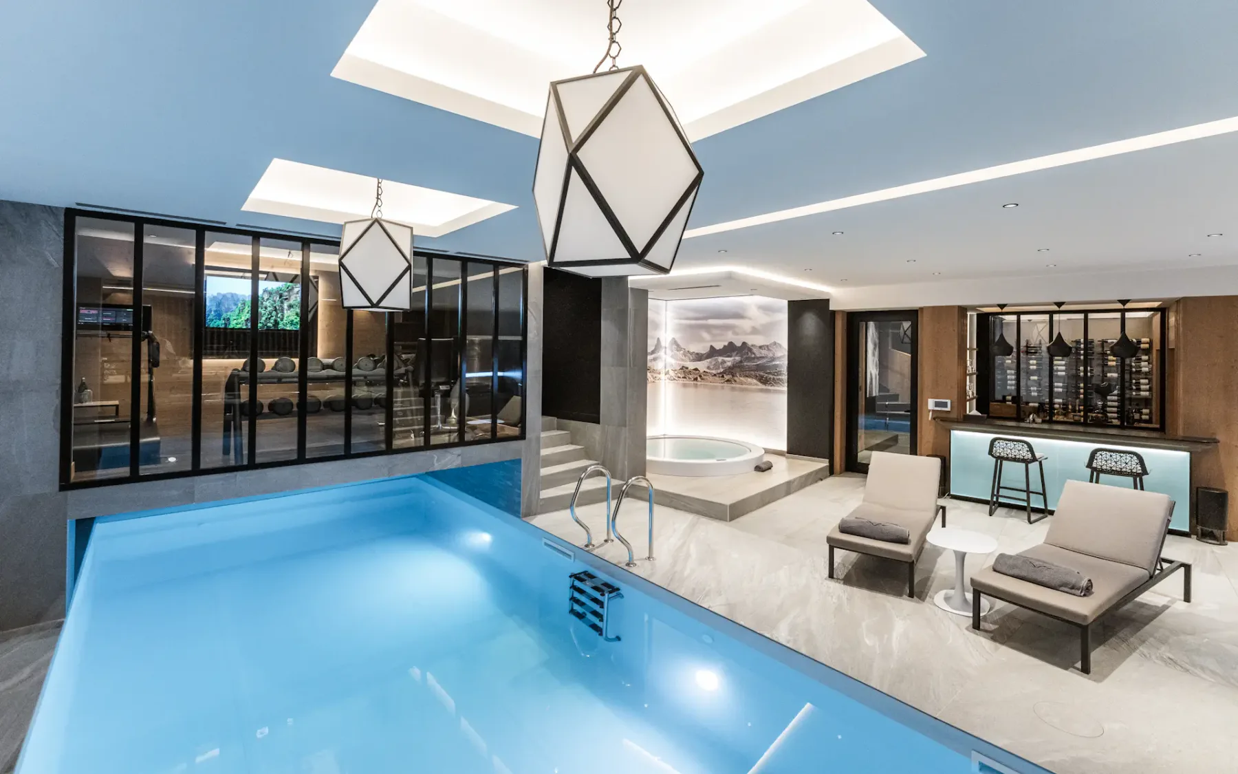 Luxurious indoor pool area with modern lighting, lounge chairs, a bar, and a hot tub, featuring sleek decor and large windows.