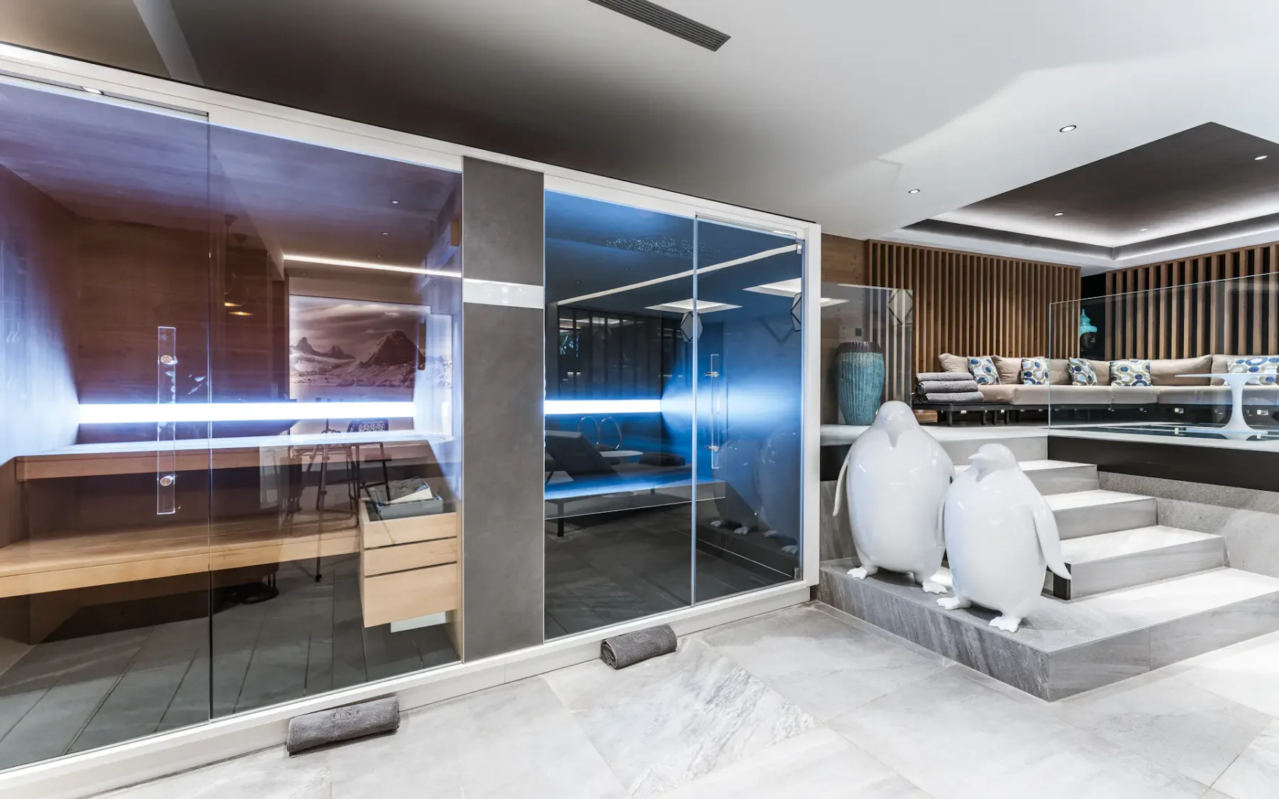 Modern spa interior with a glass-walled sauna, contemporary seating area, and decorative penguin sculptures on tiled steps.