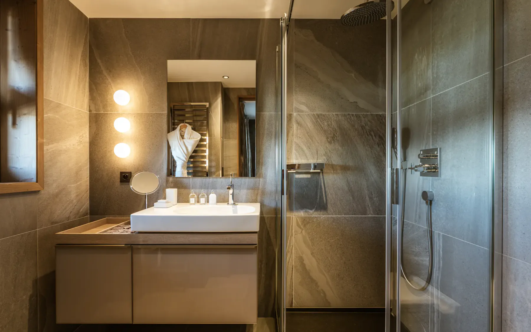 Modern bathroom with a glass shower, large mirror, illuminated vanity, and towels. Earth-toned tiles create a warm, elegant atmosphere.