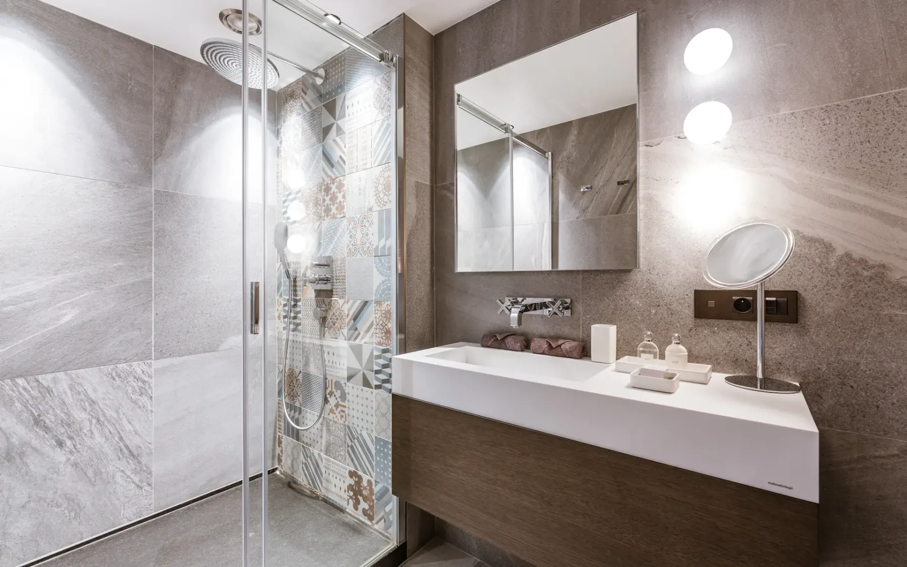 Modern bathroom with a glass shower, patterned tiles, large mirror, white sink, wooden vanity, and two round lights on the wall.