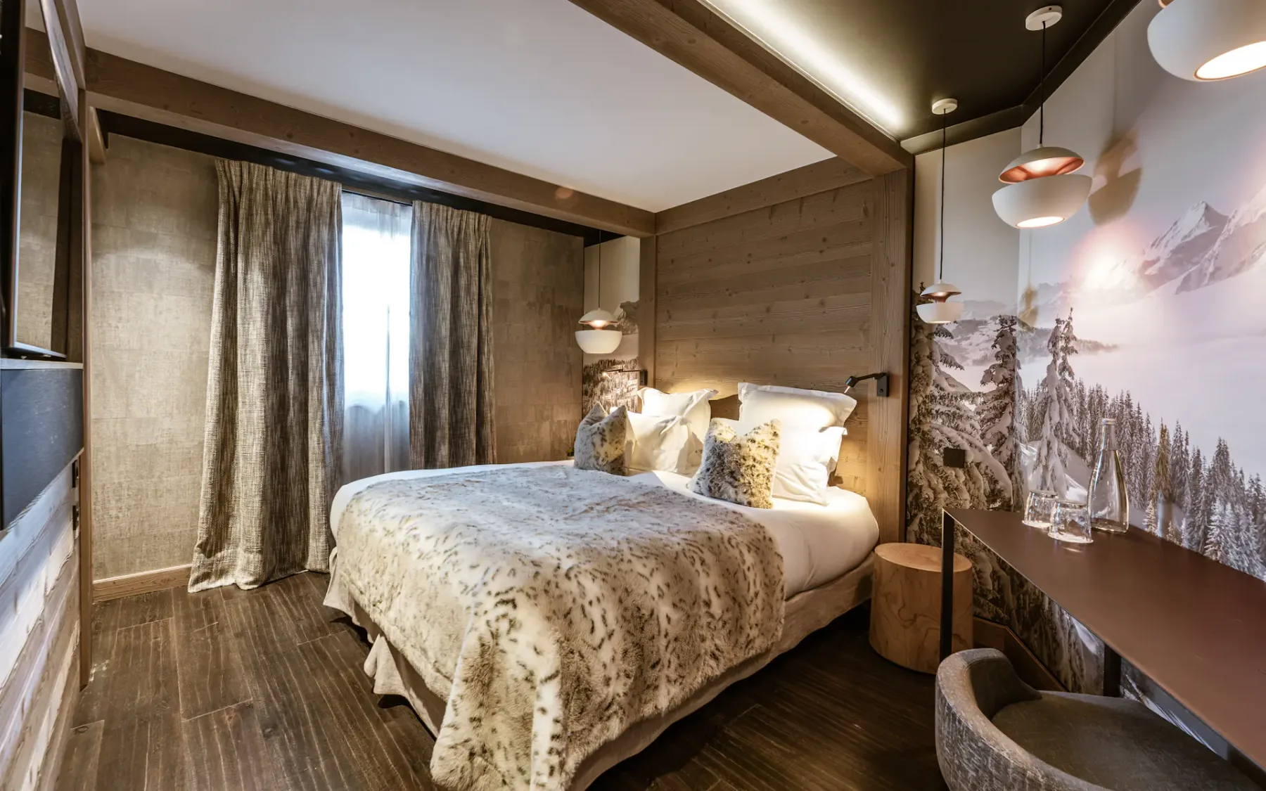 Cozy bedroom with a double bed, fur blankets, wooden walls, warm lighting, a scenic wall mural, and a small desk with a chair.