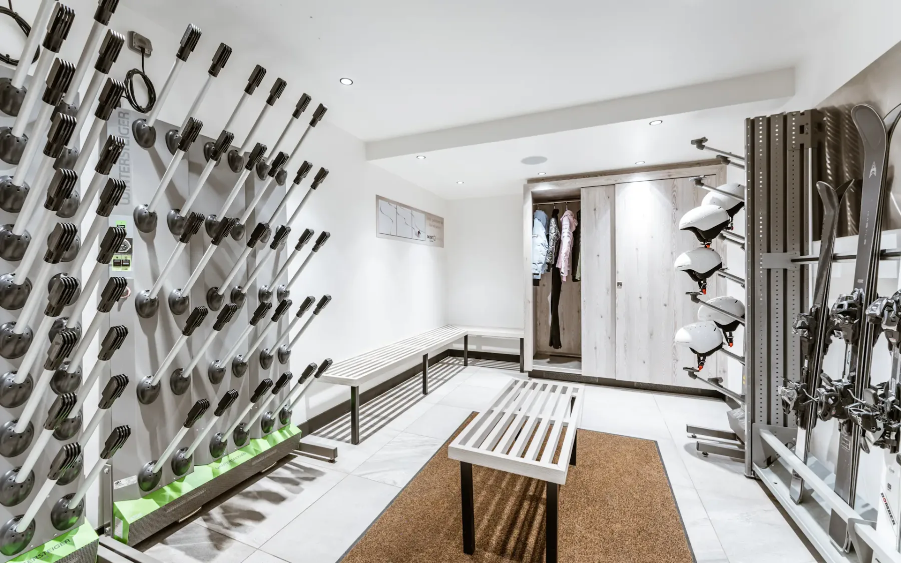 A modern ski storage room with racks holding ski poles, helmets, and boots. Benches and a brown mat are on the floor.