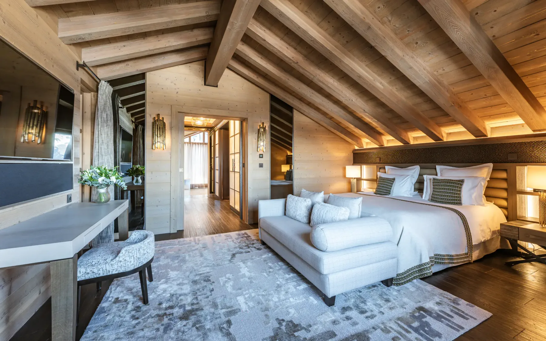 Cozy attic bedroom with wooden beams, a large bed, white sofa, desk, and soft lighting. Neutral tones create a warm, inviting atmosphere.