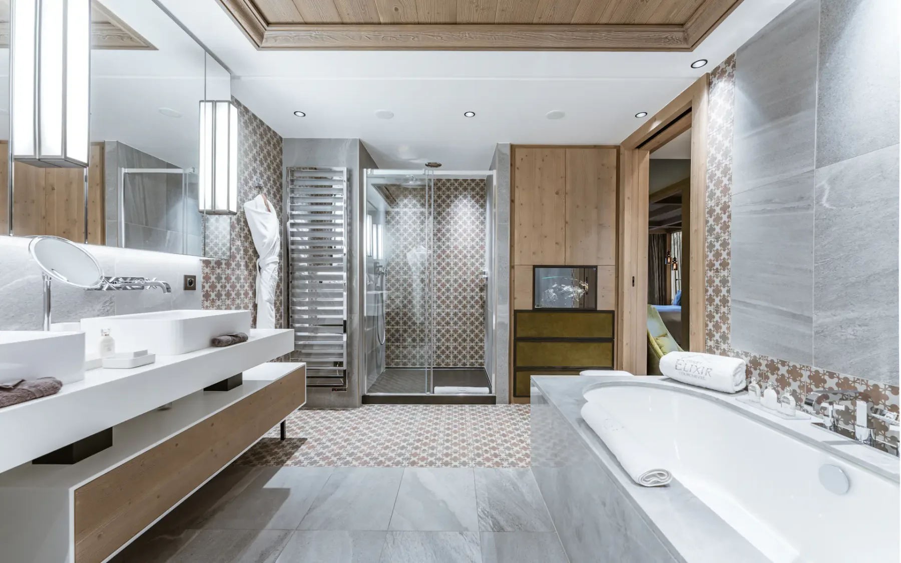 Modern bathroom with a large bathtub, dual sinks, and a glass-enclosed shower. Wooden accents and patterned tiles enhance the decor.
