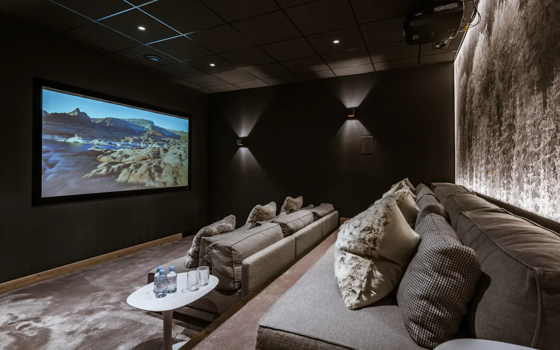 Home theater with plush gray seating, a large screen displaying a scenic landscape, and ambient lighting. Two small tables hold water bottles.