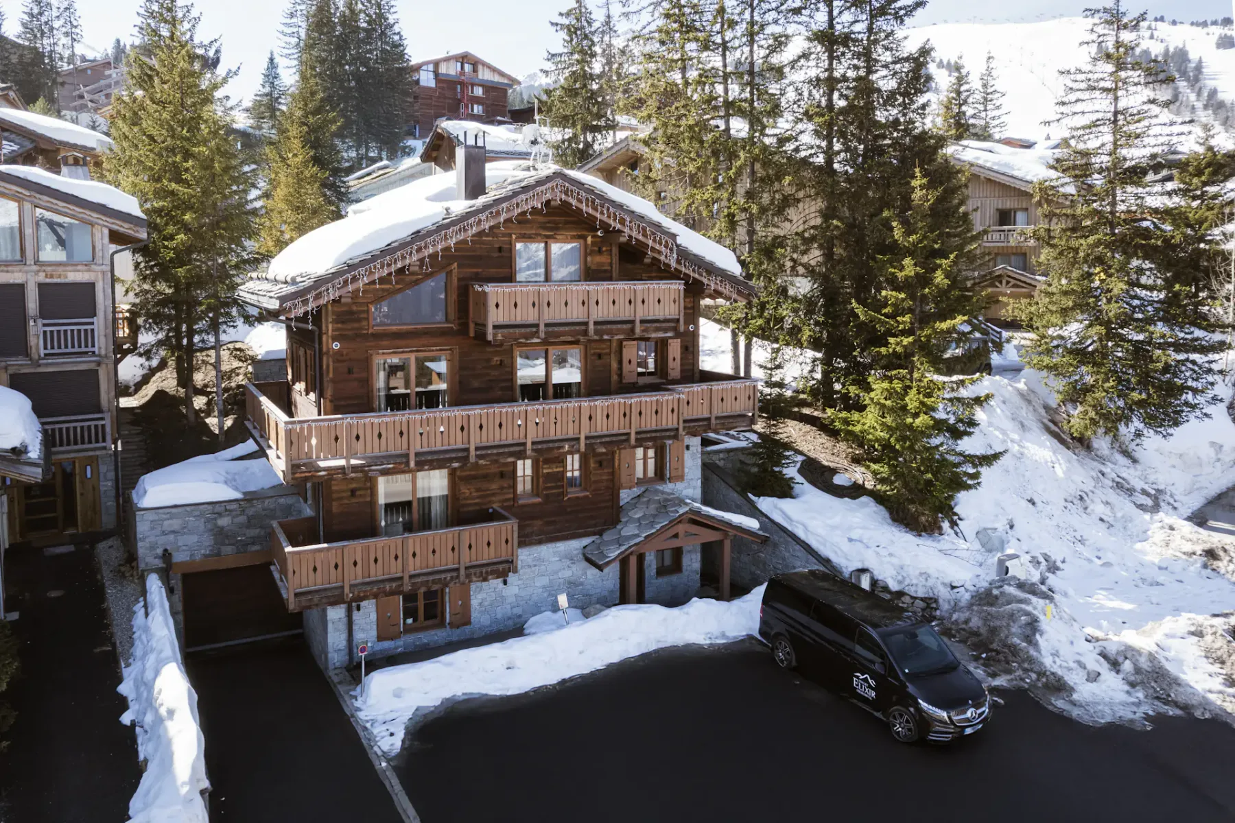 A wooden chalet with snow-covered roof and balconies, surrounded by pine trees, with a black van parked in the driveway.