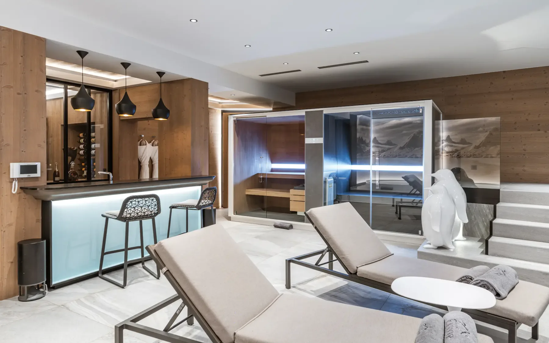 Modern spa lounge with beige loungers, a wooden bar area, sleek black bar stools, a glass-enclosed sauna, and decorative penguin sculpture.