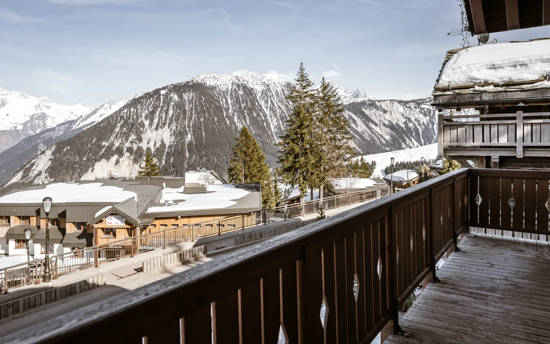 Snow-covered mountain view from a wooden balcony, overlooking a village with pine trees under a clear sky.