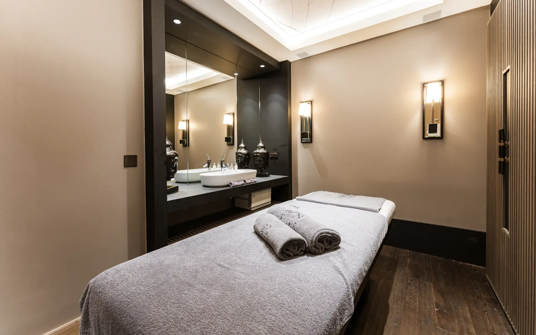A serene spa room with a massage table, neatly folded towels, soft lighting, and a mirror above a sink.