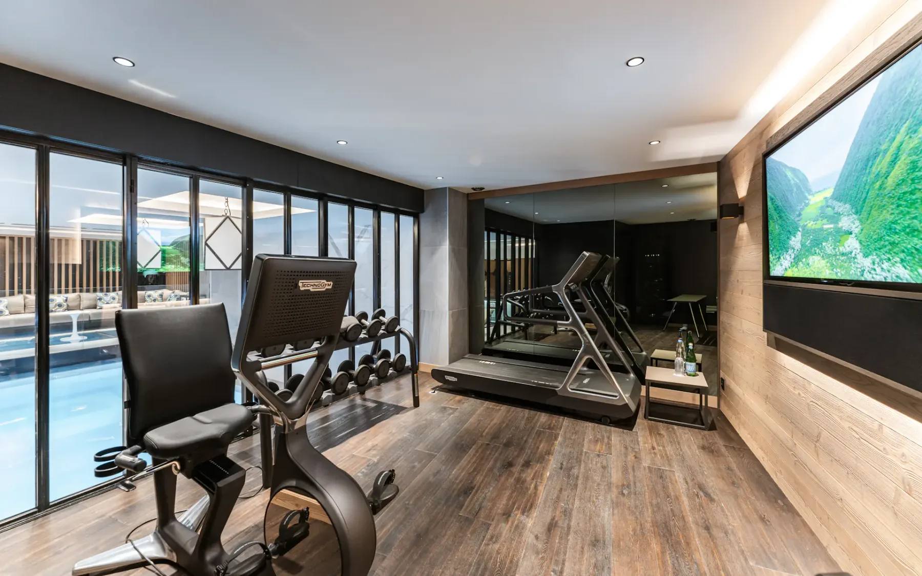 Modern home gym with exercise bike, treadmill, dumbbells, large mirror, and wall-mounted TV. Glass doors reveal a view of an indoor pool.